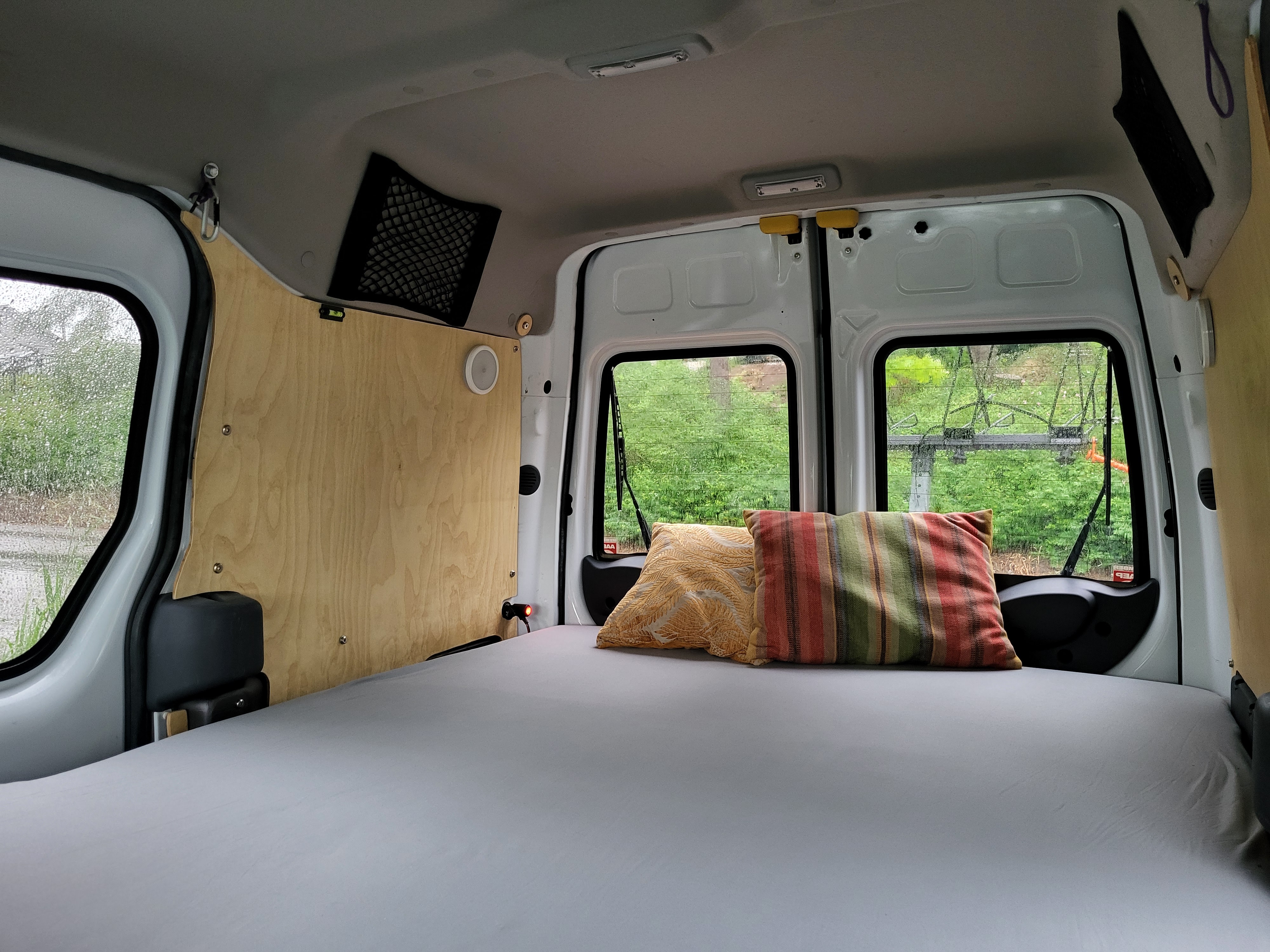 RV rental interior: A comfy bed with colorful pillows in a camper van, overlooking lush greenery through the back windows.. Ford Transit 2013