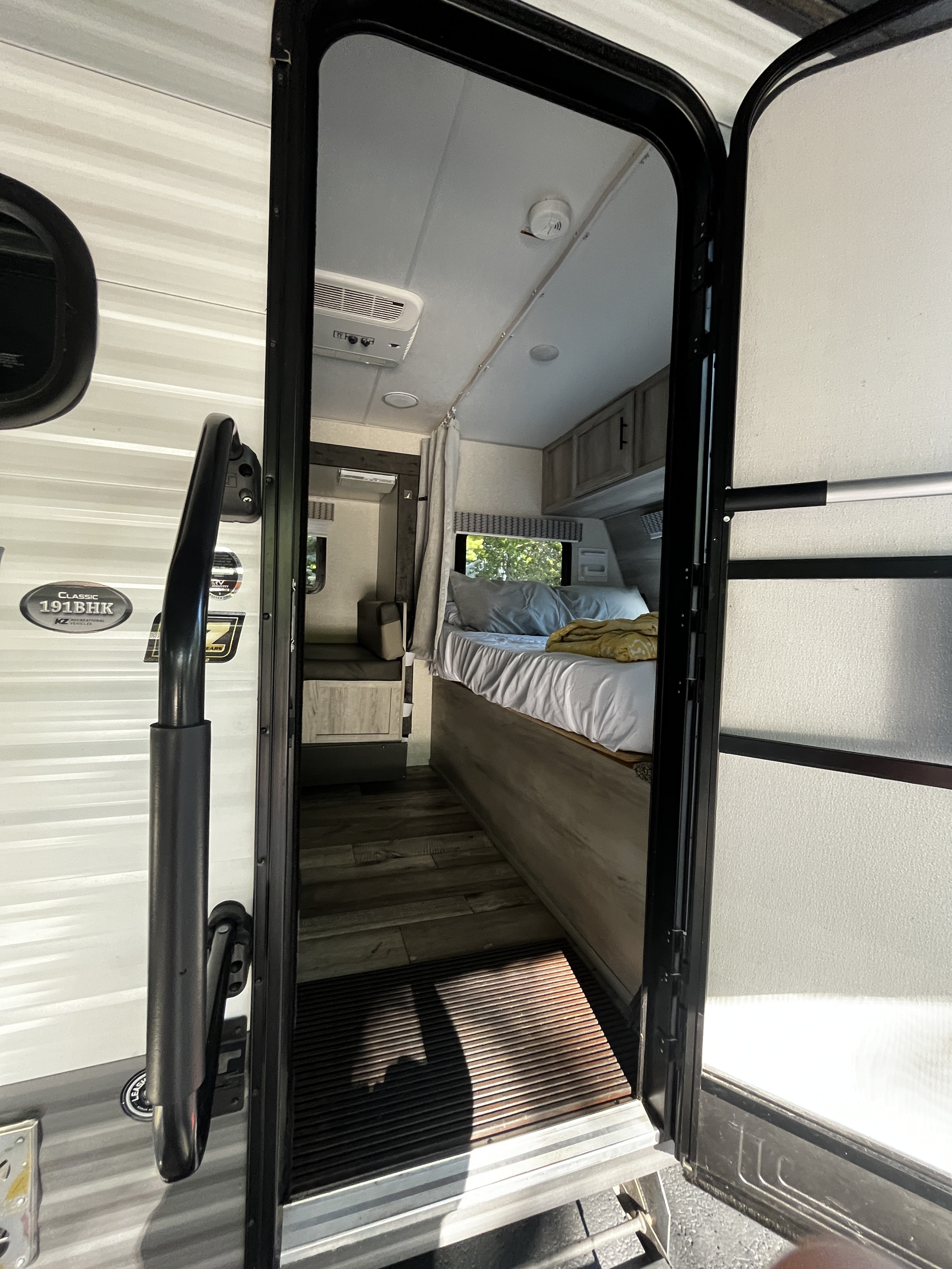 View from the open door of an RV rental, revealing a cozy bedroom, AC, wood floors, and the camper's exterior siding.. KZ Sportmans Classic 191bhk 2021