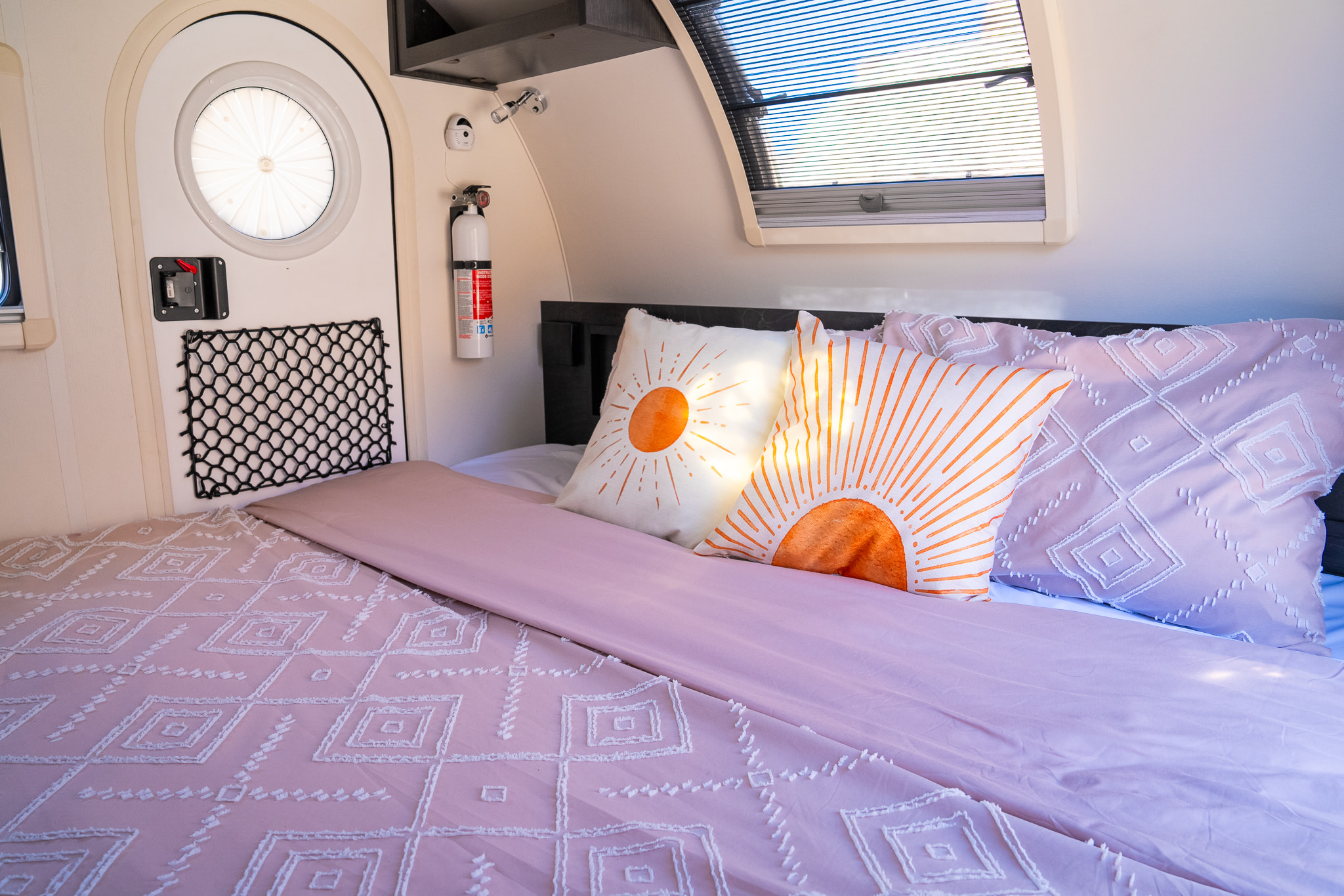 RV rental bedroom featuring a comfy bed with pink bedding, vibrant sun pillows, a round door, and a safety extinguisher.. nuCamp T@G 2023