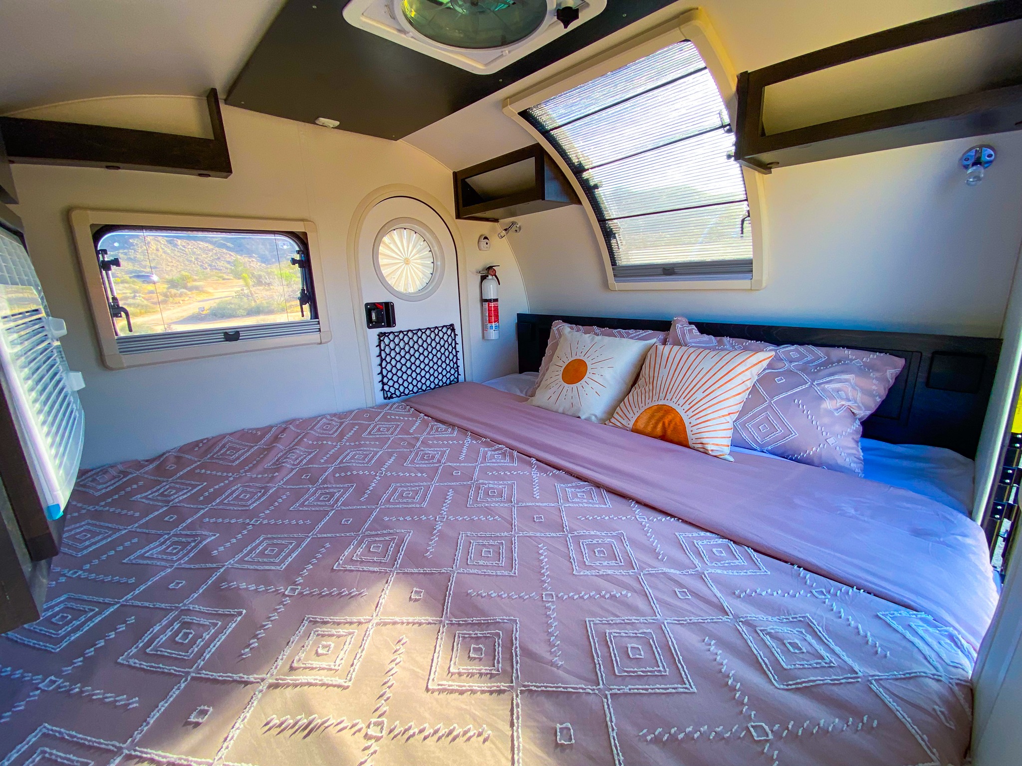 Inside an RV rental: a cozy bedroom with pink geometric bedding, scenic windows, and overhead storage for travel comfort.. nuCamp T@G 2023