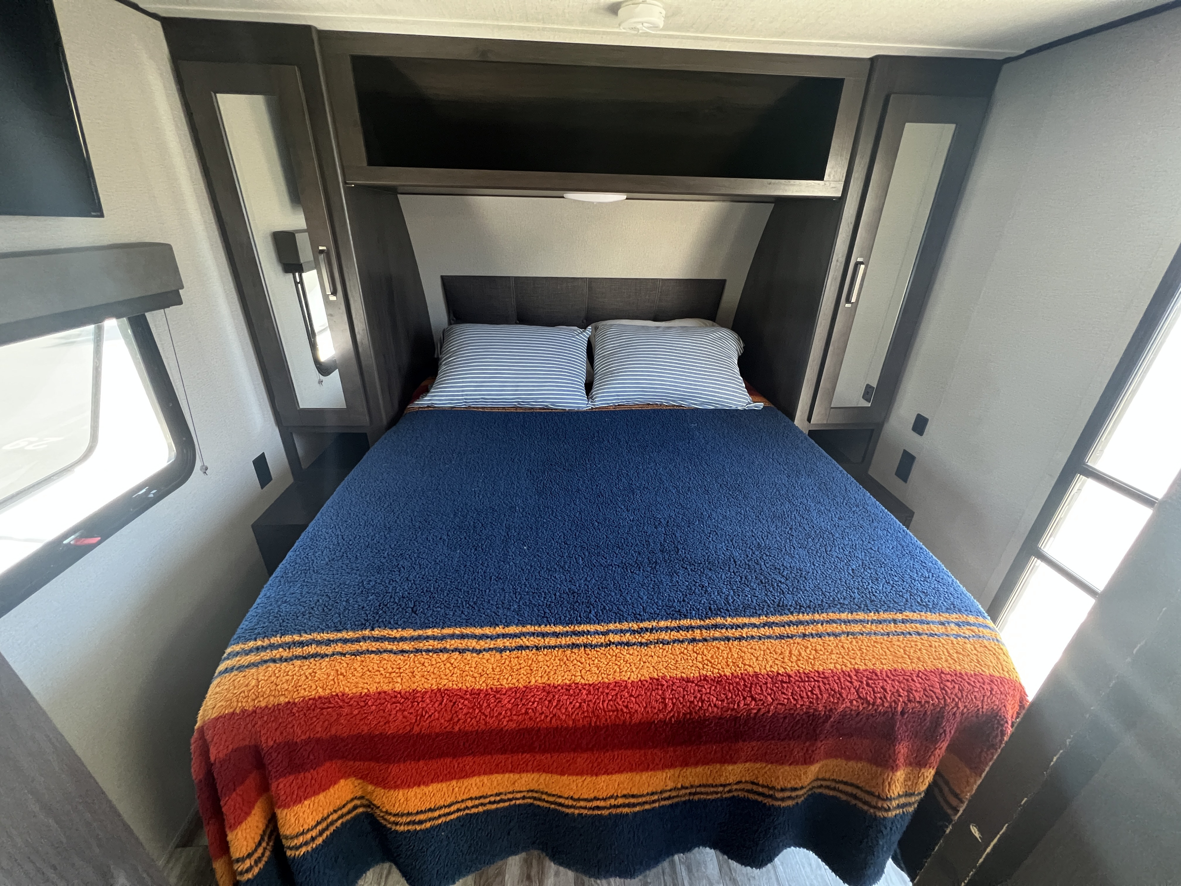 Cozy RV rental bedroom featuring a comfortable bed with striped bedding, mirrored cabinets, and side windows.. Grand Design Transcend Xplor 2021