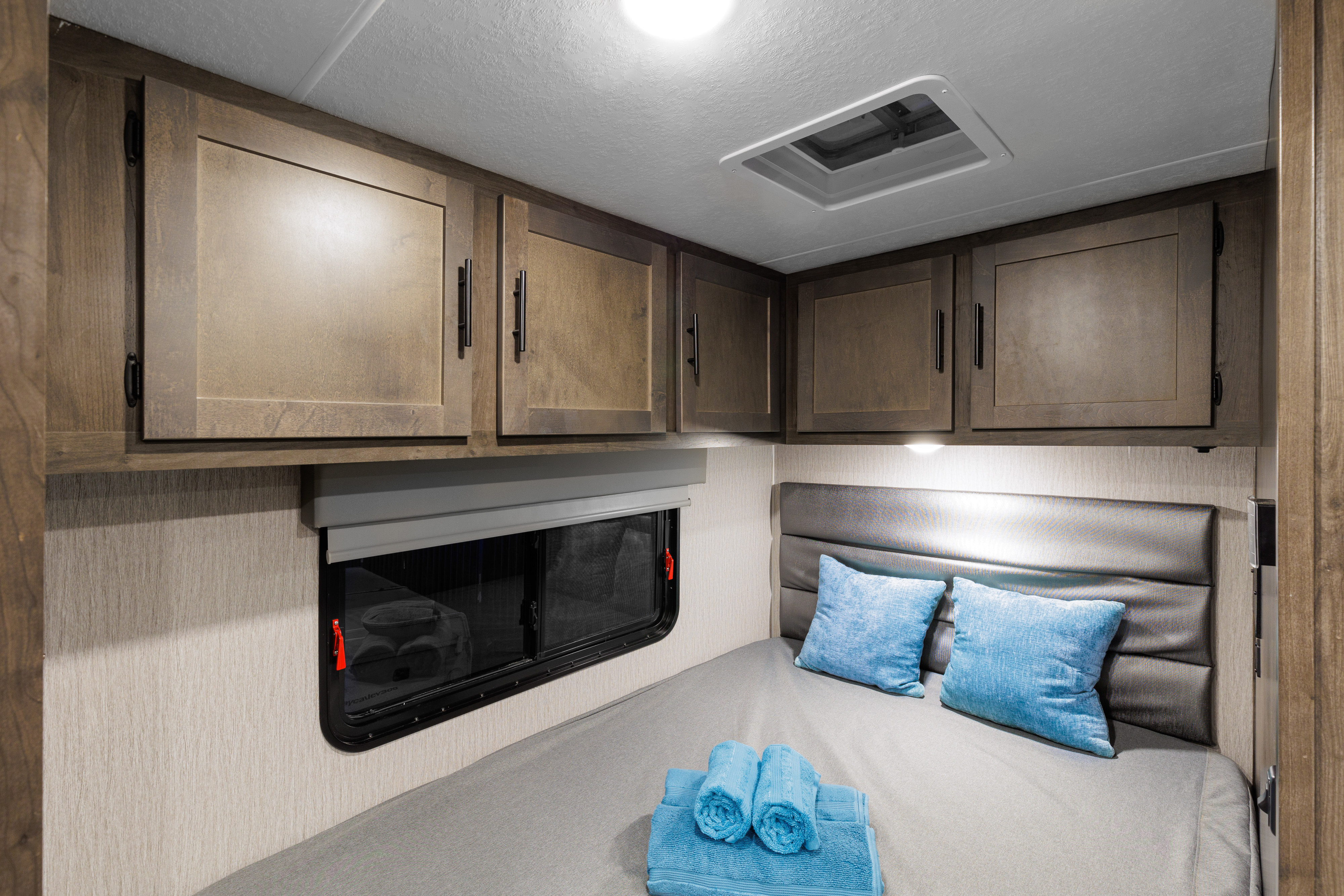An RV rental bedroom showcasing a comfortable bed with blue pillows and towels, plus wooden overhead storage cabinets.. Coachmen Prism 2023