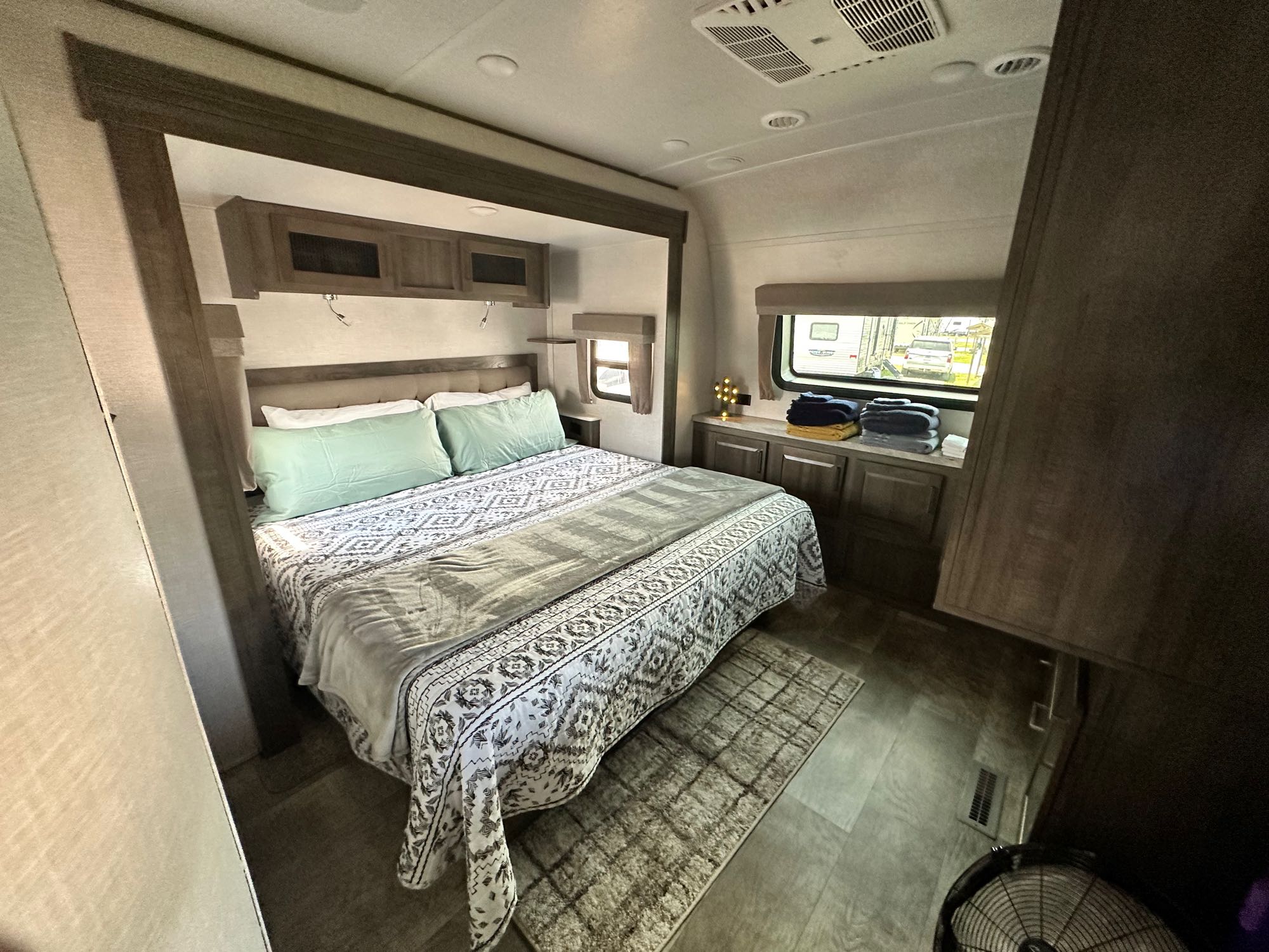 Inside an RV rental bedroom showing a made bed with green pillows, patterned quilt, built-in cabinets, and large windows.. Forest River Flagstaff Classic 2022