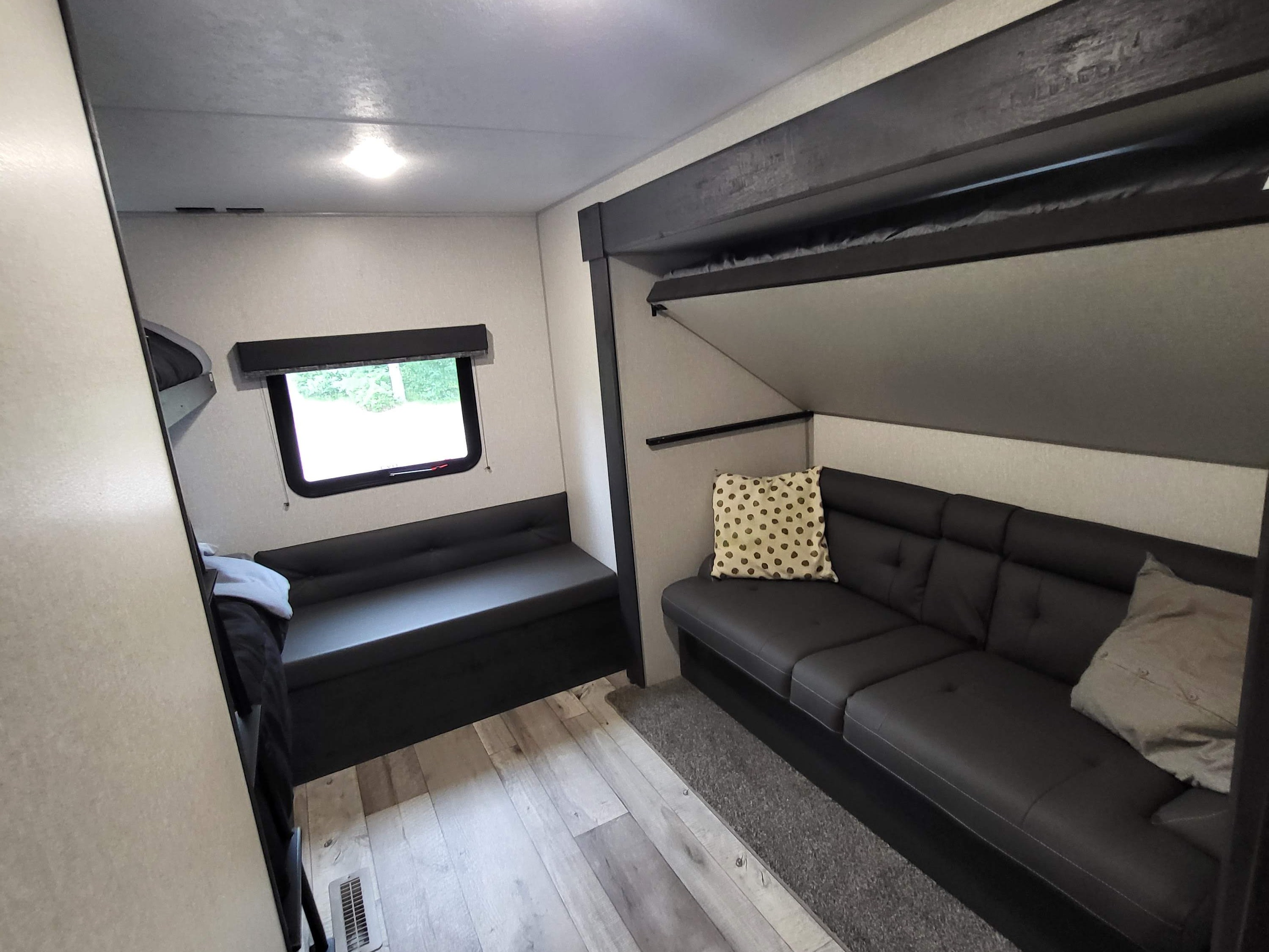 Bright RV rental interior with fold-up bunk beds, a dark L-shaped sofa, and bench seating by a window.. Sportsmen SE 2022