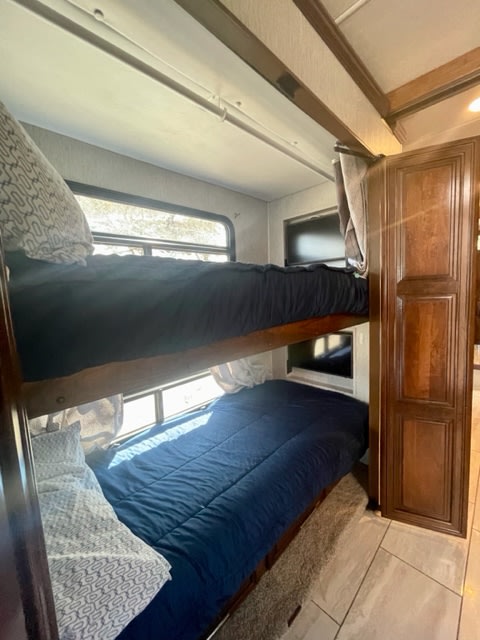 Inside a modern RV rental: a comfortable two-tier bunk bed setup with windows, bedding, and adjacent wooden cabinetry.. Forest River Berkshire 2021