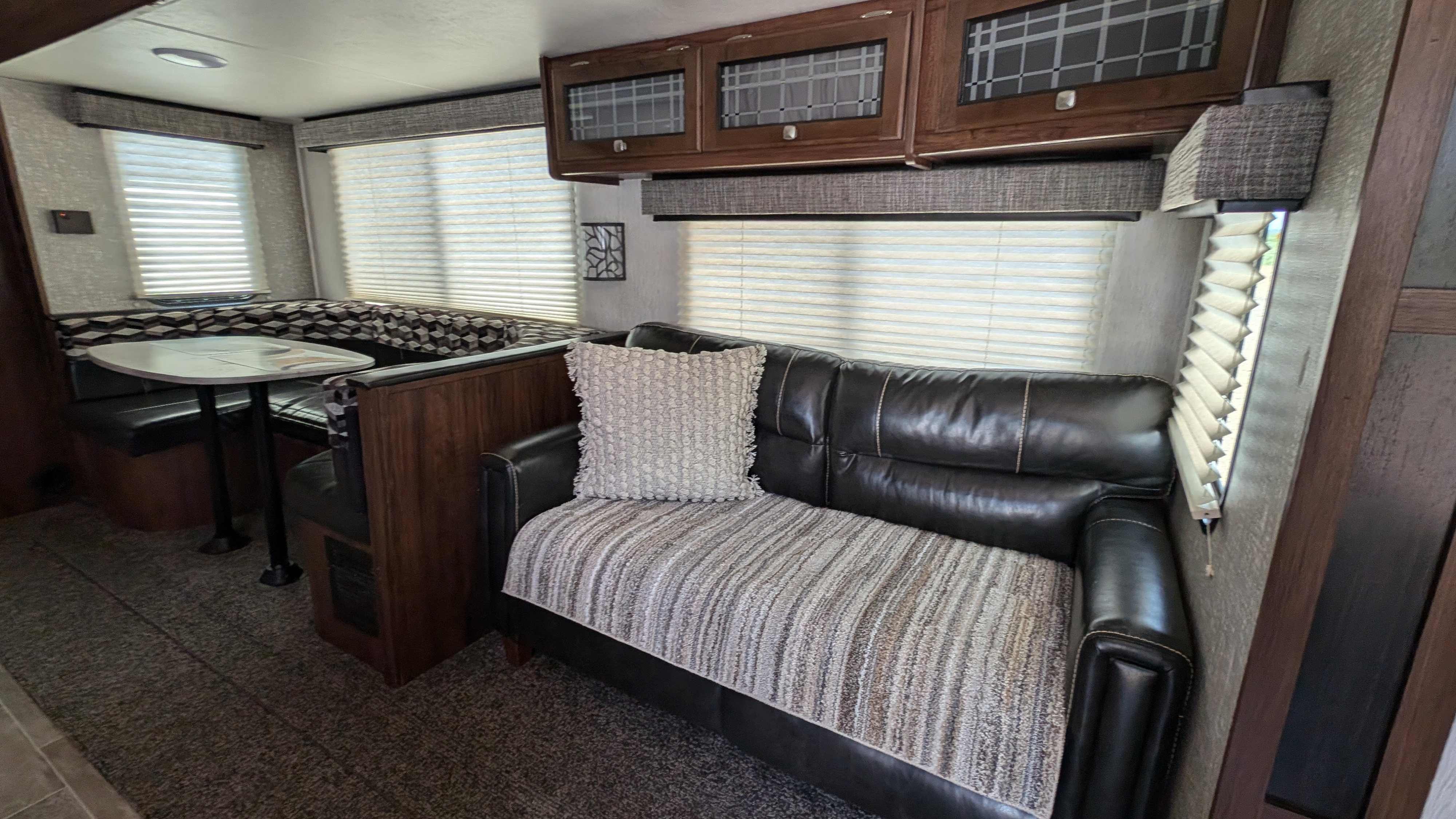 Inside this RV rental: black leather sofa with throw, dining booth, overhead cabinets, & blinds for a functional living space.. Heartland RVs Mallard 2019