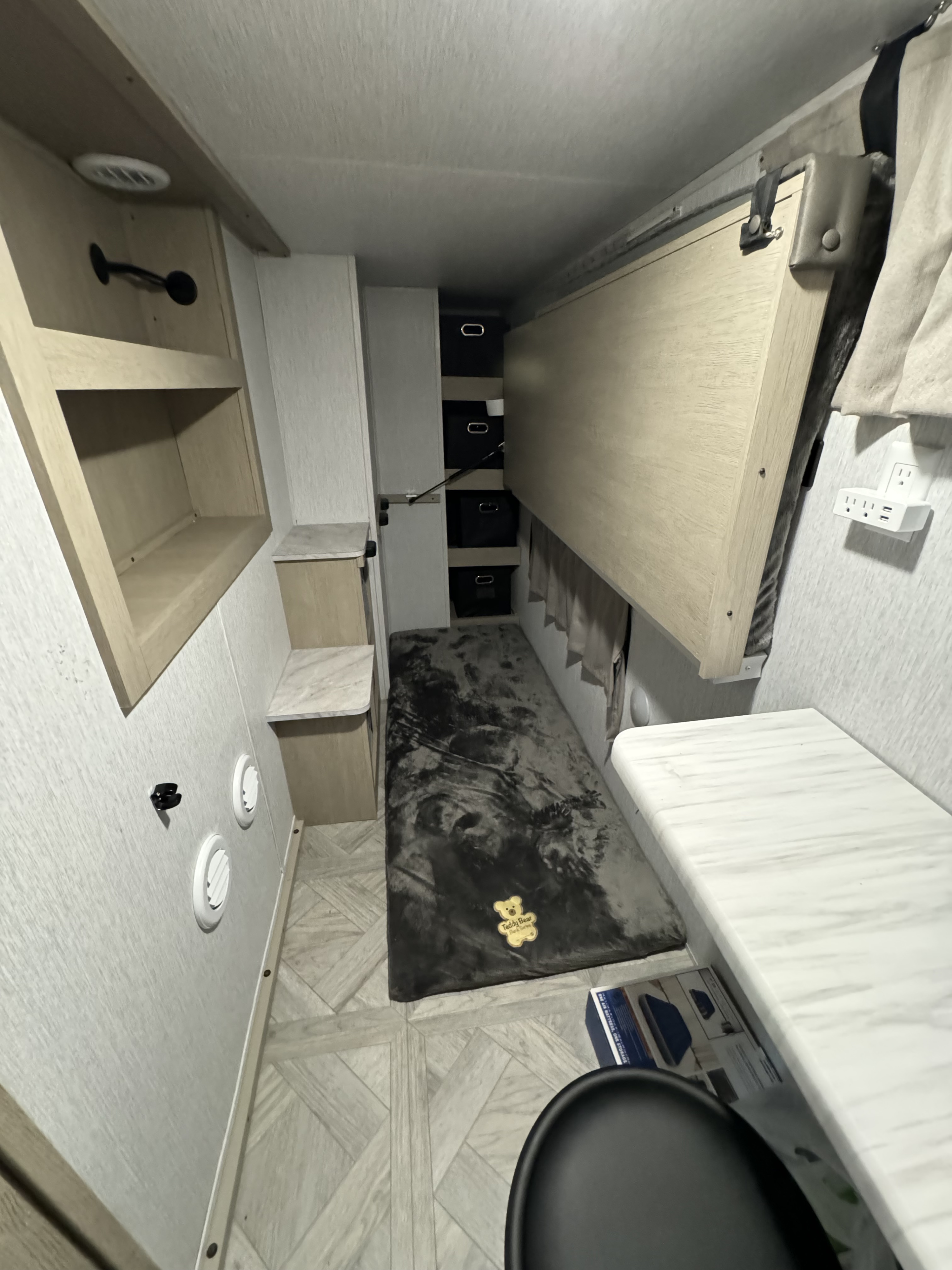 Compact RV rental interior featuring a wall-mounted folding bed, shelves, and a gray rug, providing functional living.. Forest River Wildwood Platinum 2024
