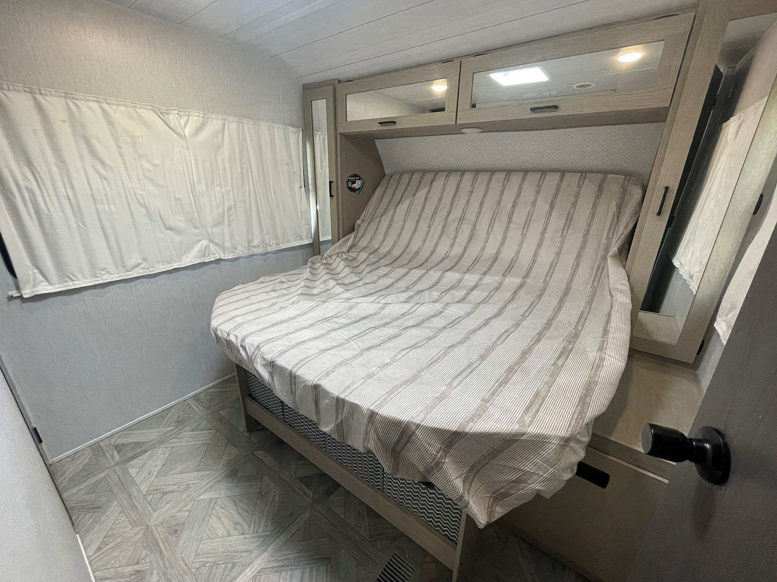 Light-toned RV rental bedroom: a striped bed under mirrored overhead cabinets, with a window shade.. Forest River Wildwood 2022