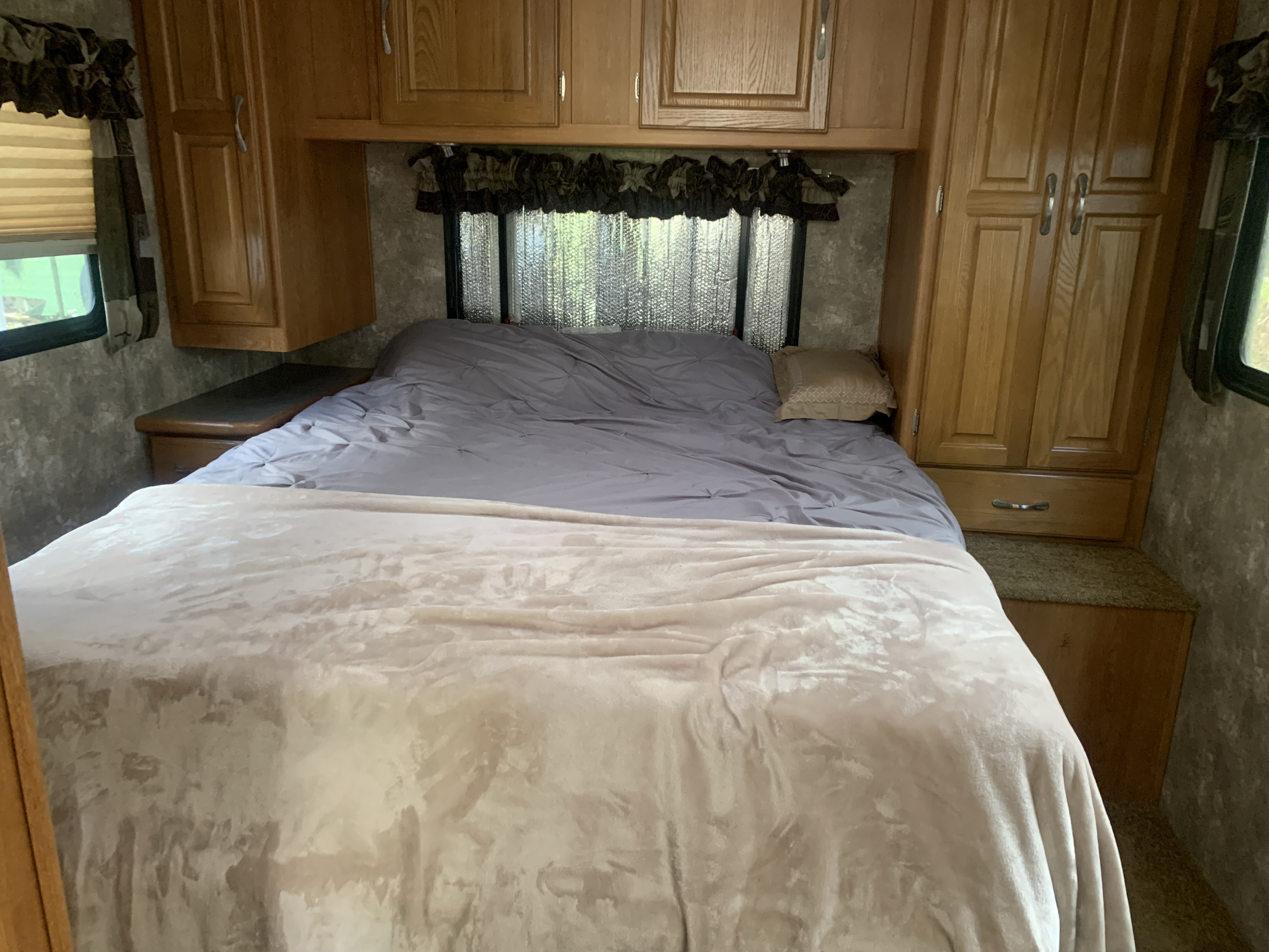 This inviting RV rental bedroom boasts a bed with grey and light blankets, wood cabinets, and outdoor views.. Coachmen Freelander 2007