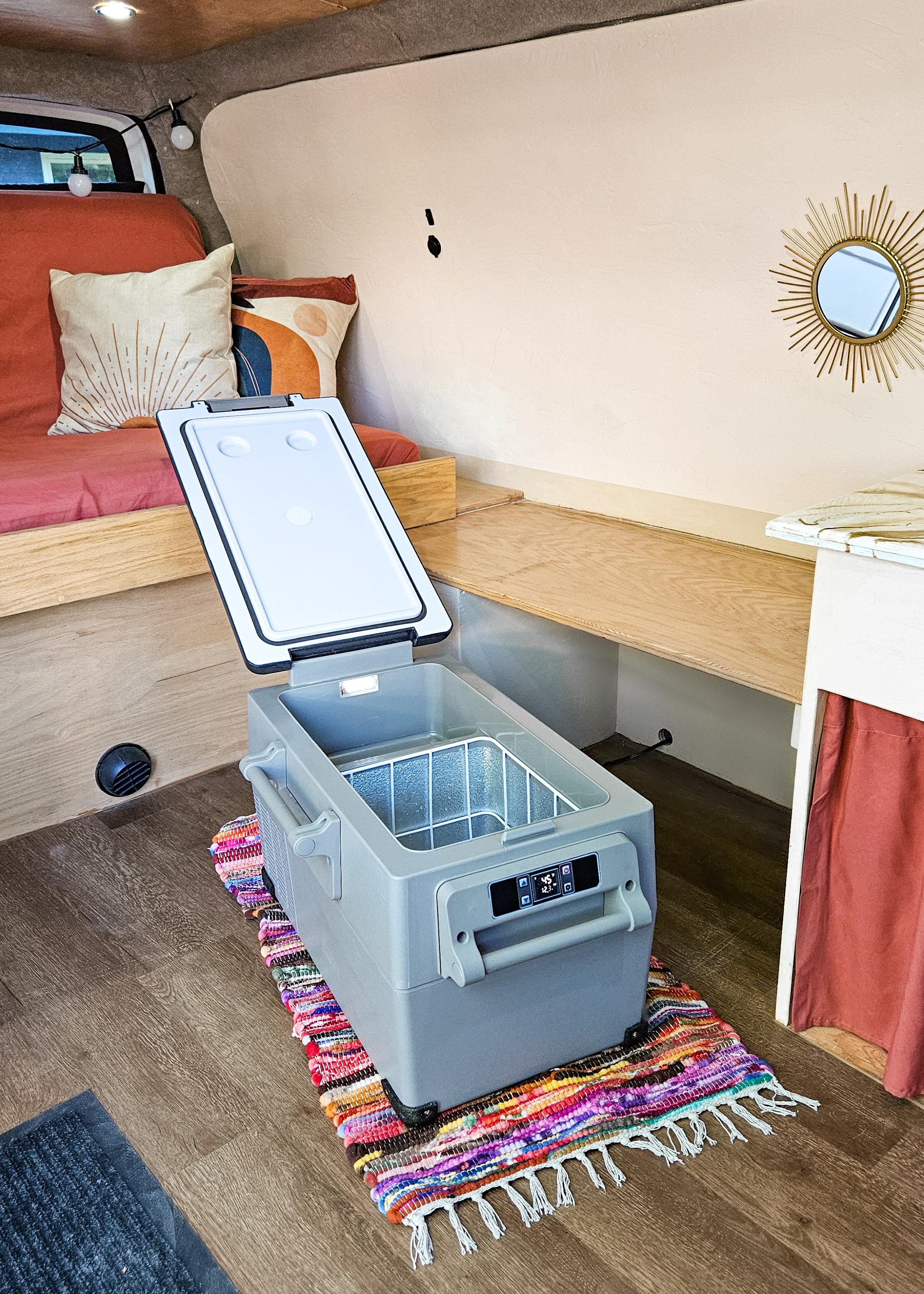 Spacious RV rental interior featuring a portable fridge on a colorful rug, wooden bed, and shelving for a comfy road trip setup.. Gmc Savanna 2003