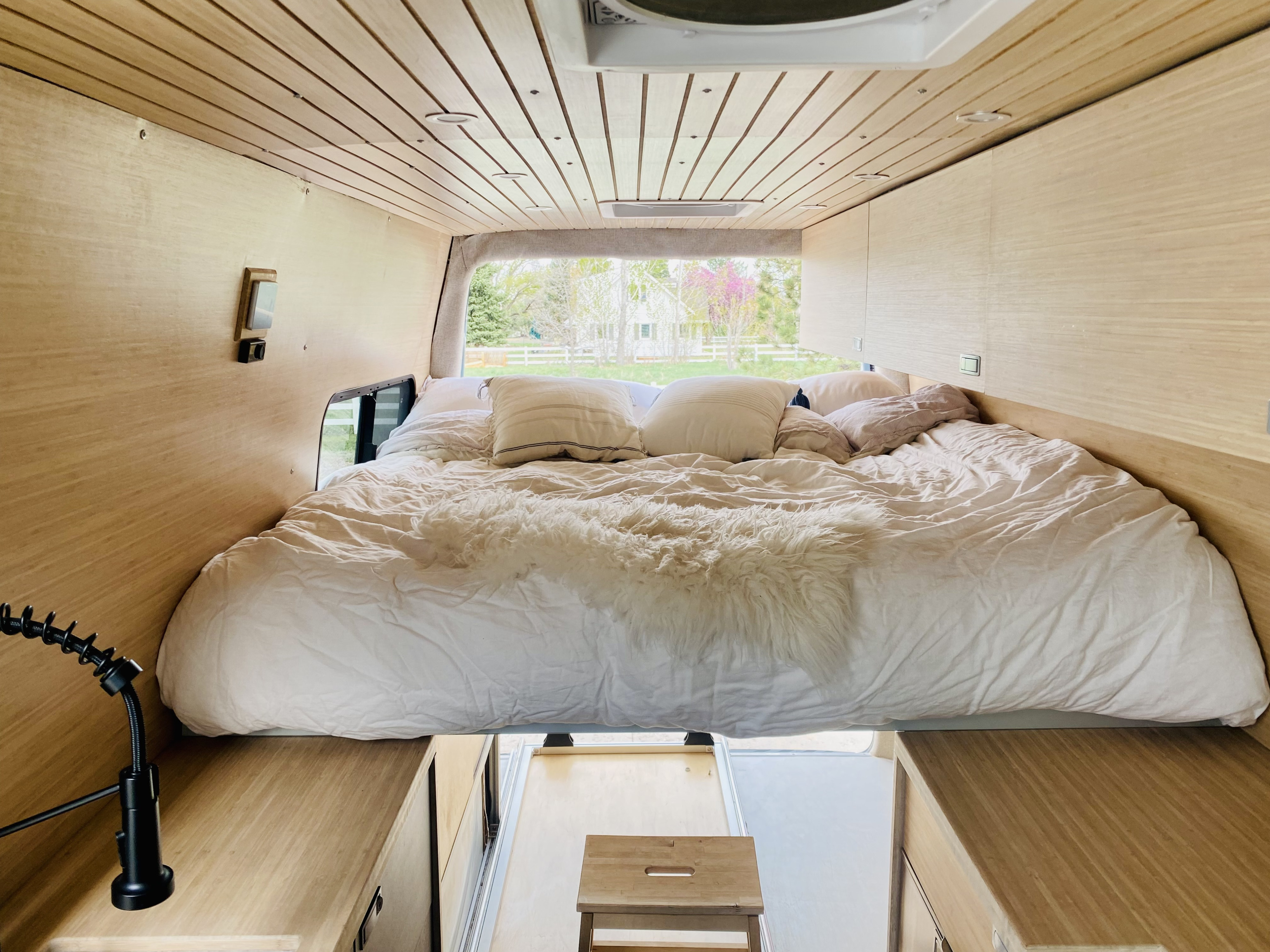 Interior of an RV rental camper van showing a large bed with white bedding, light wood walls, and a view outside.. Mercedes Sprinter 2500 2020