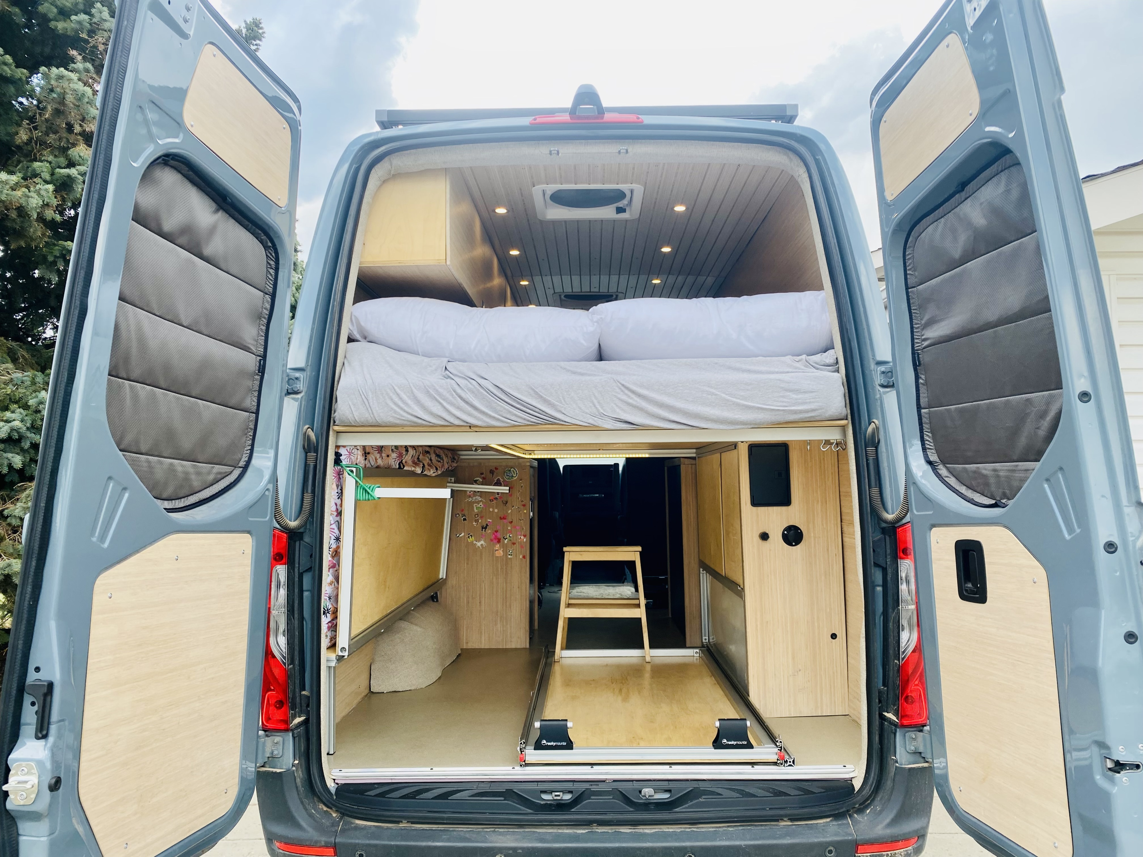Rear view of a spacious camper van interior featuring a cozy upper bed, pull-out storage, and wood finishes, ideal for an RV rental.. Mercedes Sprinter 2500 2020