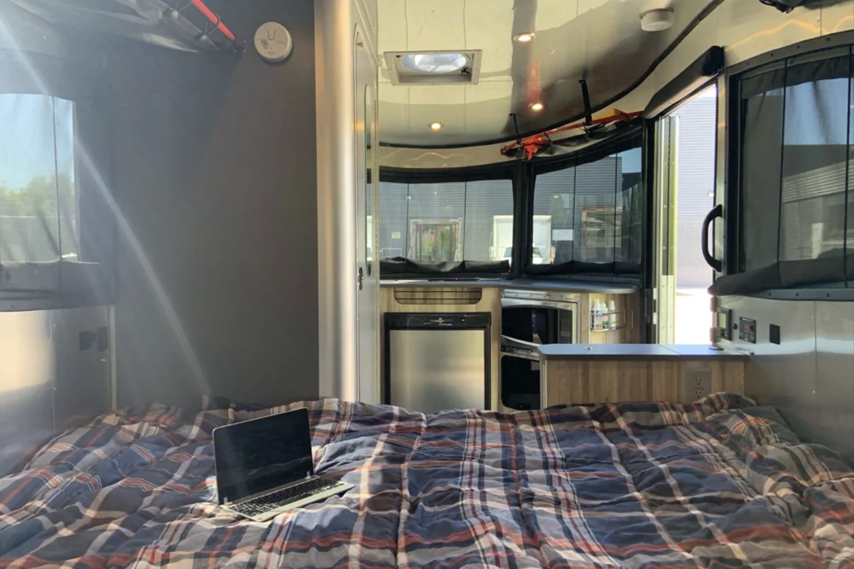 View inside an RV rental showing a plaid bed with laptop, compact kitchen with fridge and microwave. Cozy travel space.. Airstream Basecamp 2018