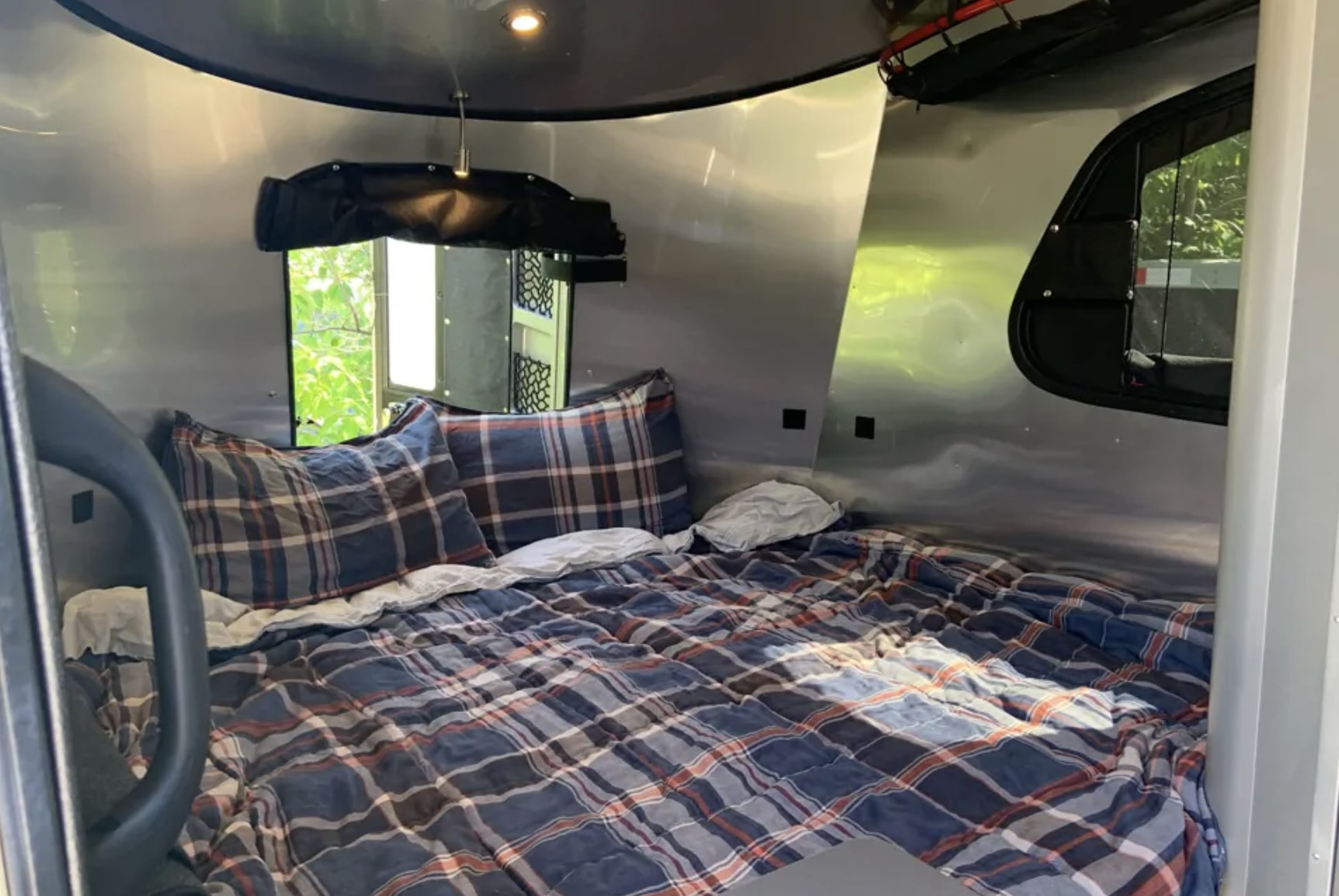 Plush plaid bed inside a sleek rv rental. Polished aluminum walls and large windows reveal green outdoor scenery.. Airstream Basecamp 2018