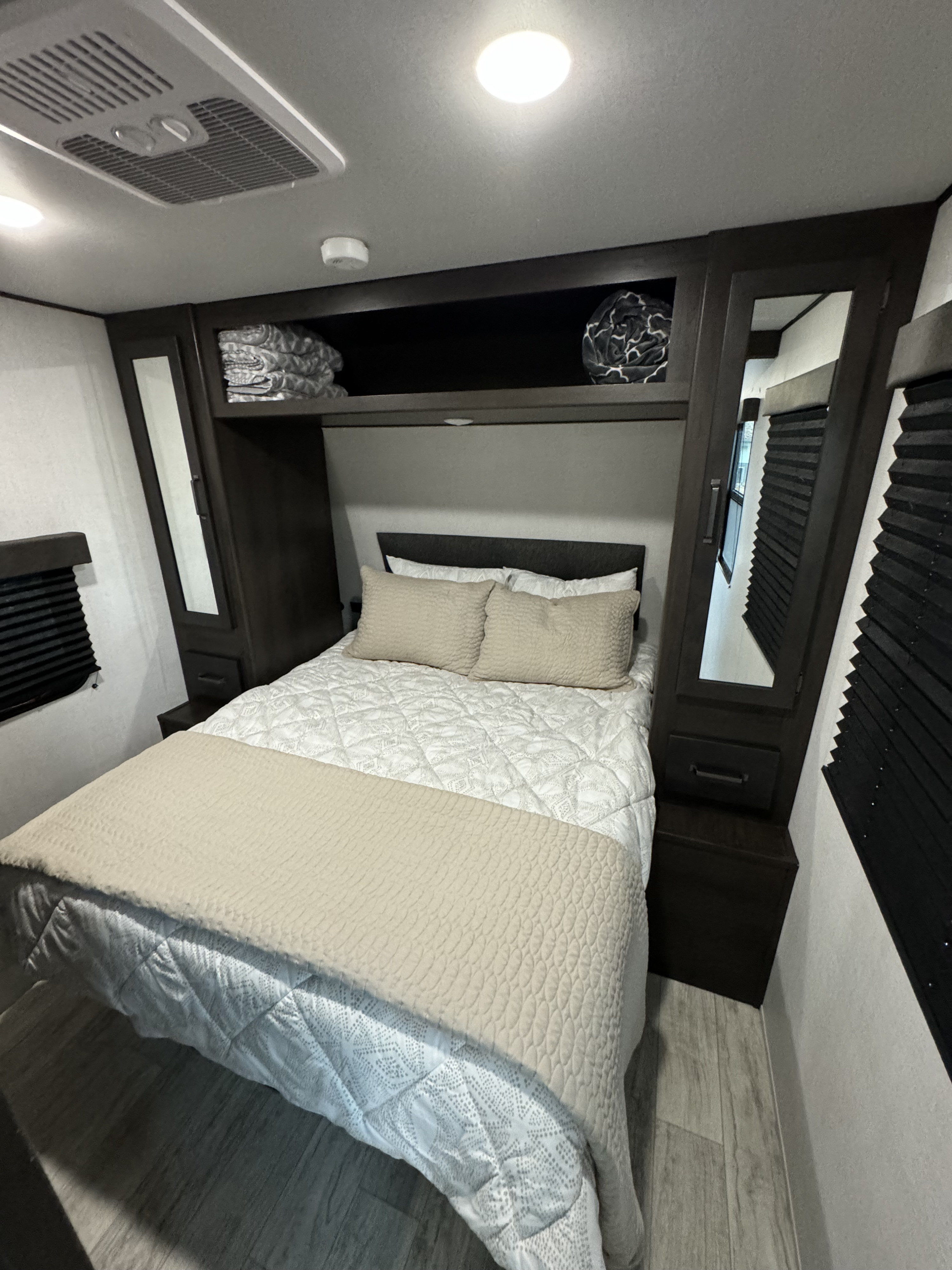 Cozy RV rental bedroom featuring a neutral-toned bed, dark wood built-in storage, and bright overhead lighting.. Grand Design 27MAV 2024