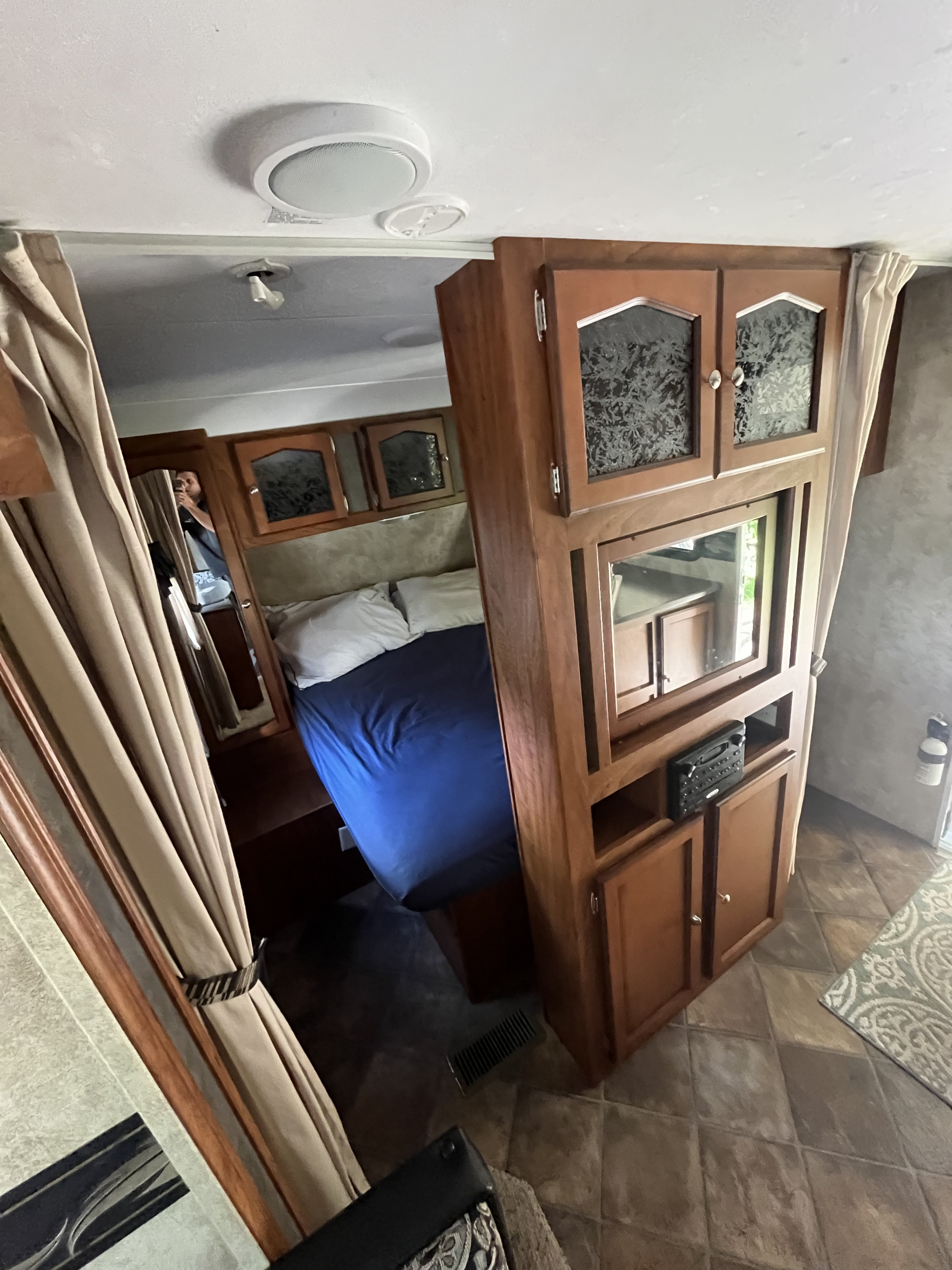 Inside an RV rental: a compact bedroom with a bed, overhead cabinets, and a central storage/entertainment unit.. Keystone RV Passport Ultra 2013
