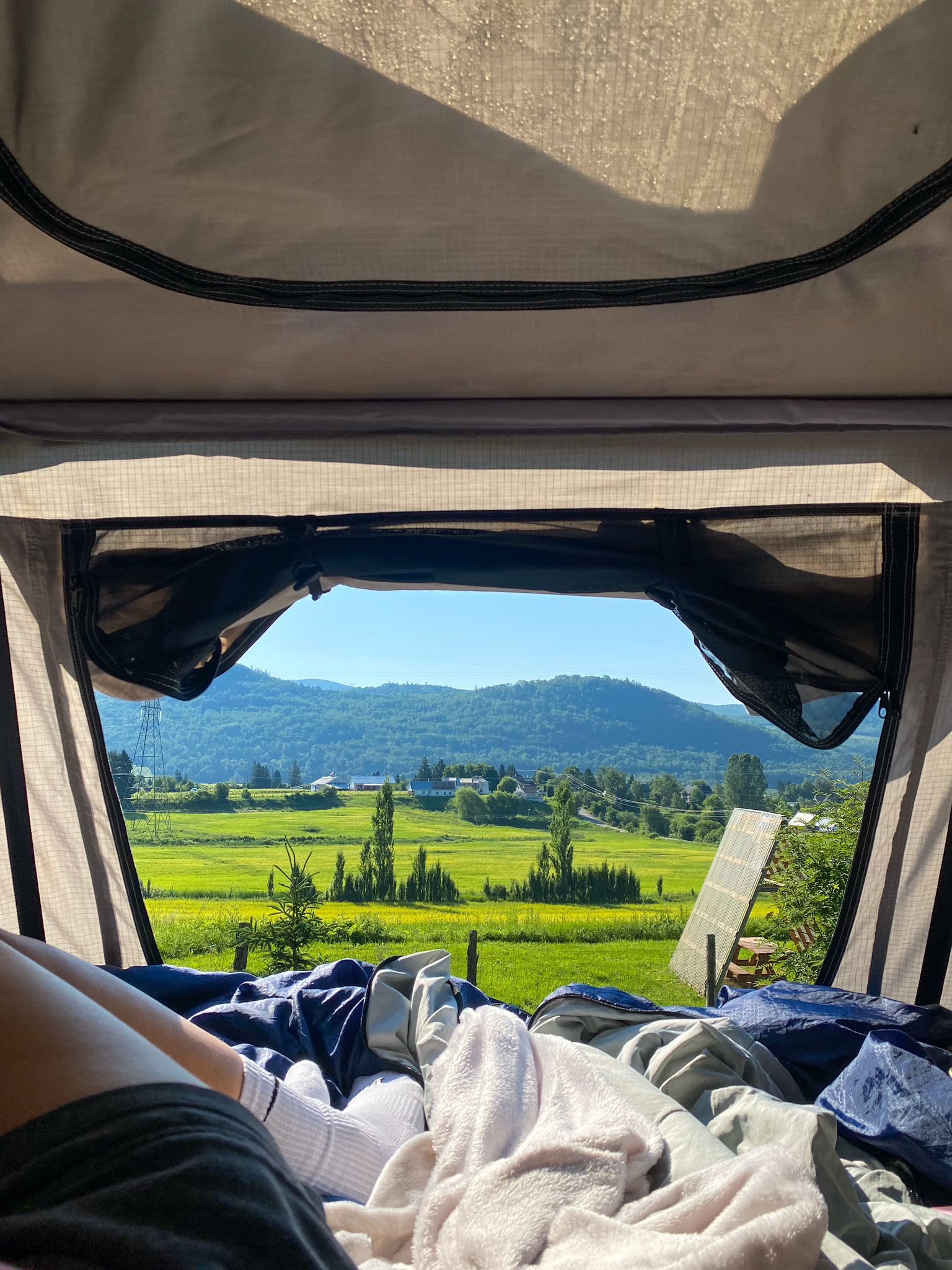 Waking up to a serene view from an RV rental. Inside: legs & blankets. Outside: green fields, mountains, and a peaceful valley.. Ford Escaper 2013