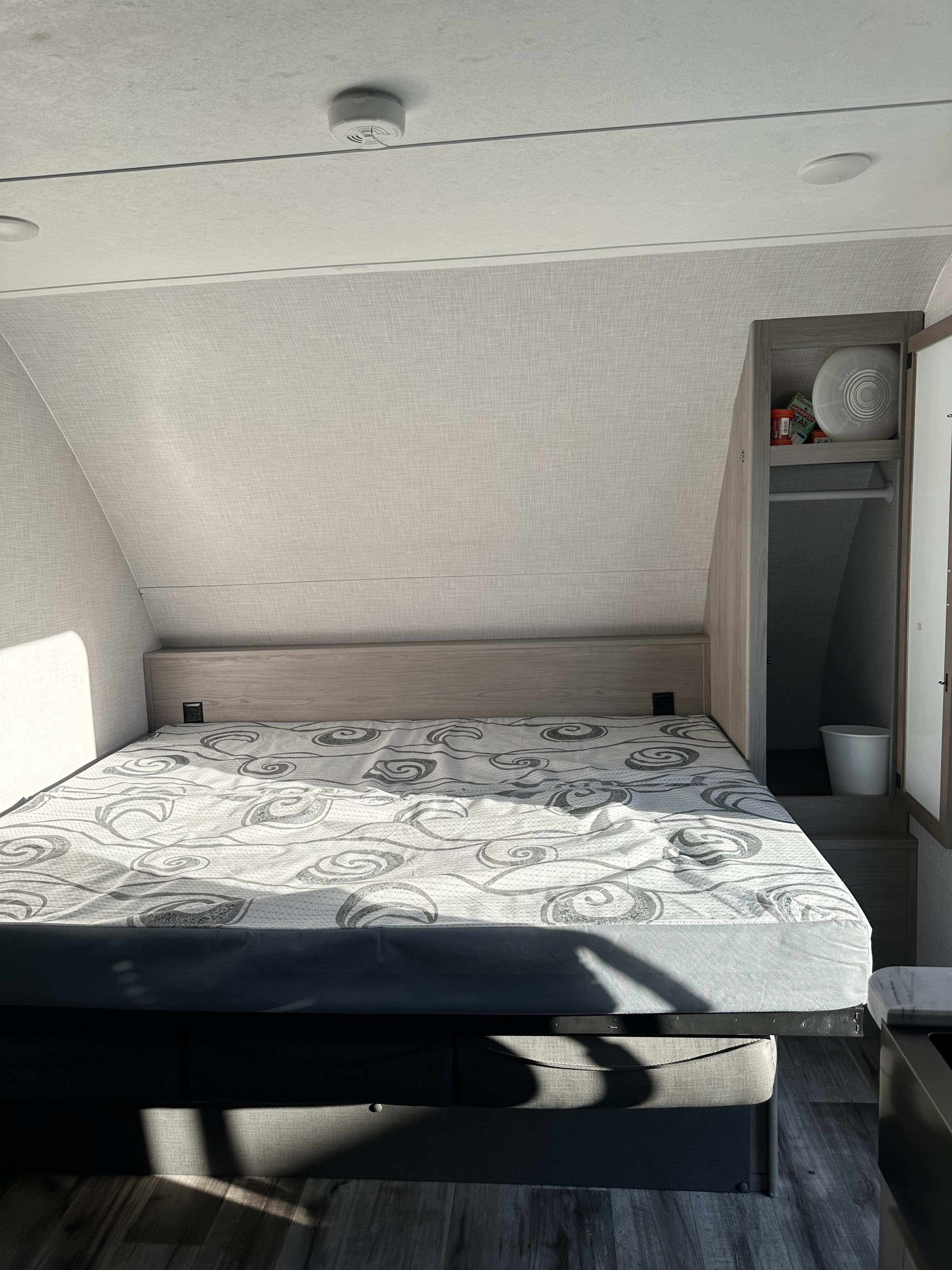 RV rental bedroom: A bed with a grey swirly mattress, light wood headboard, and storage cabinet with games. Sunlight visible.. Dutchmen Coleman Light 2022