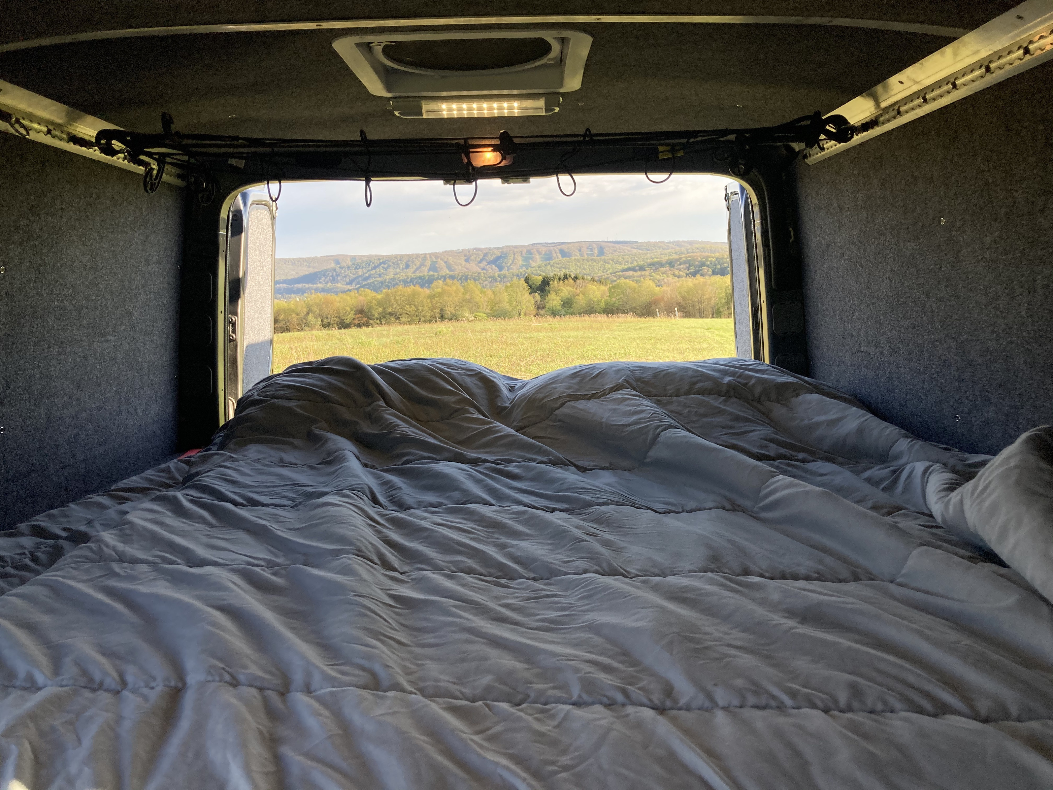Cozy rv rental interior with a comfortable bed facing open back doors, revealing a scenic field and distant green mountains.. Mercedes-Benz Sprinter 2016