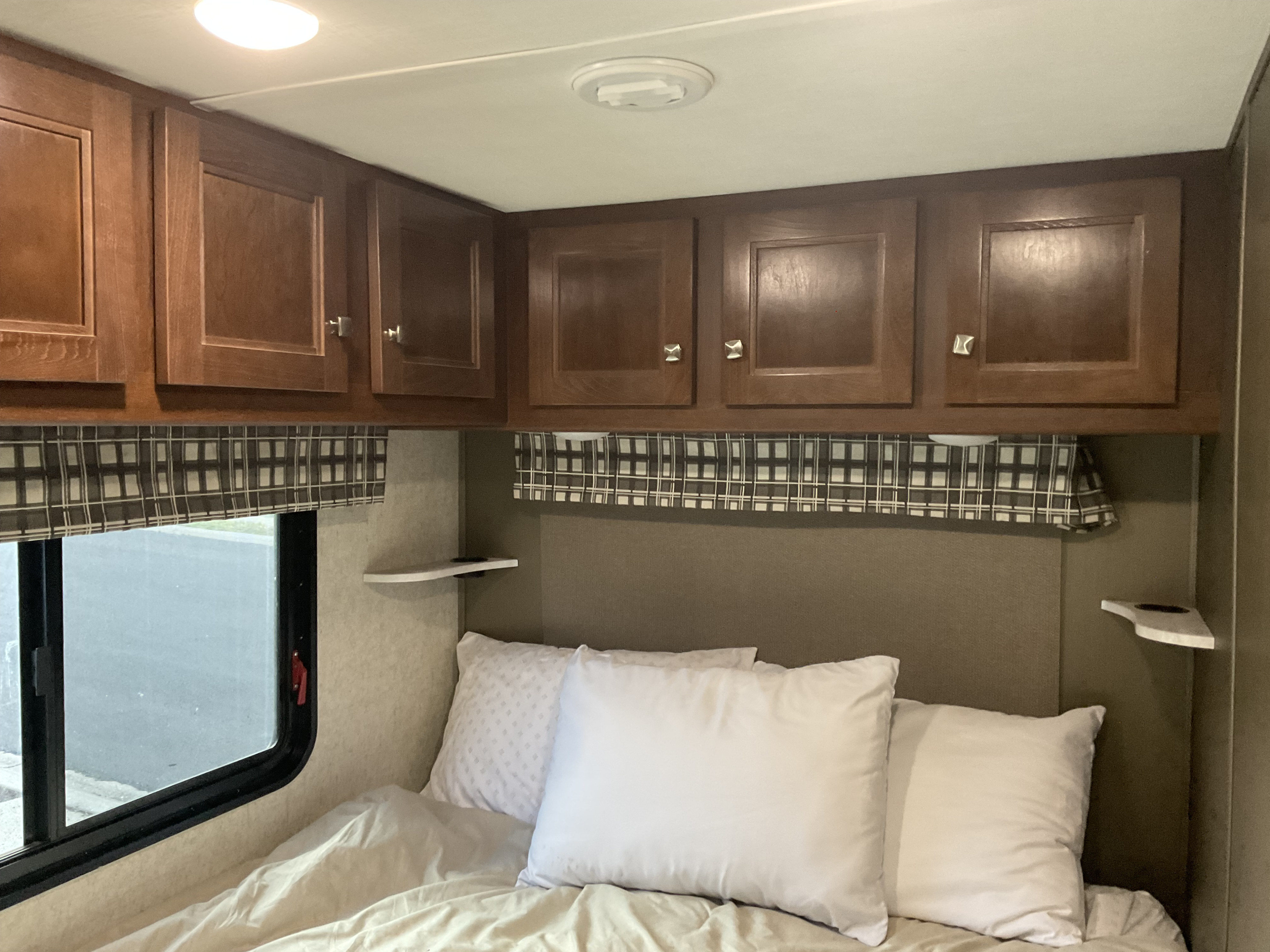 View of an RV rental bedroom with dark wood cabinets, a window, and a bed with white pillows and neutral bedding.. Forest River Sunseeker 2019