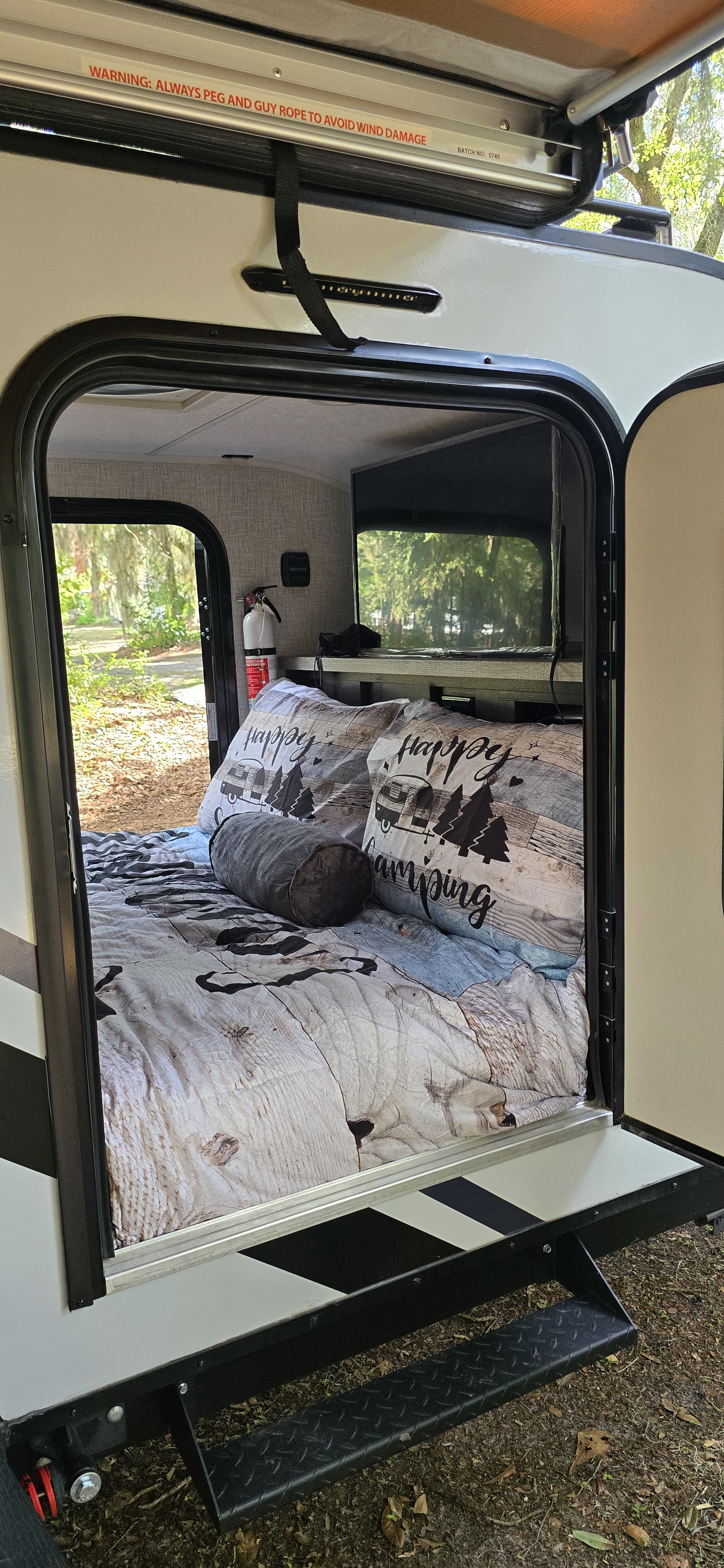 An inviting RV rental interior shows a bed with "Happy Camping" decor, TV, and a fire extinguisher.. Braxton Creek Bushwhacker 2024
