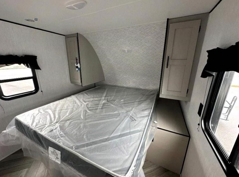 This RV rental bedroom interior features a new mattress in plastic, flanked by overhead cabinets and windows, ready for a cozy trip.. Heartland RVs Prowler Lynx 2023