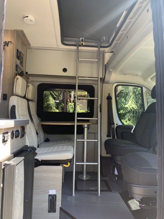 This cozy camper van interior is perfect for your next RV rental adventure, featuring a dining area, bed, and ladder to an upper bunk for mobile living.. Winnebago Solis 2022
