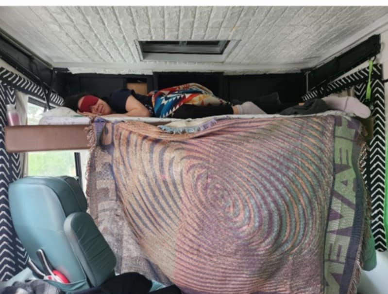 Inside an RV rental, someone naps on an elevated bed with an eye mask. A child's car seat sits below. Cozy travel.. Safari Trek 2610 1994