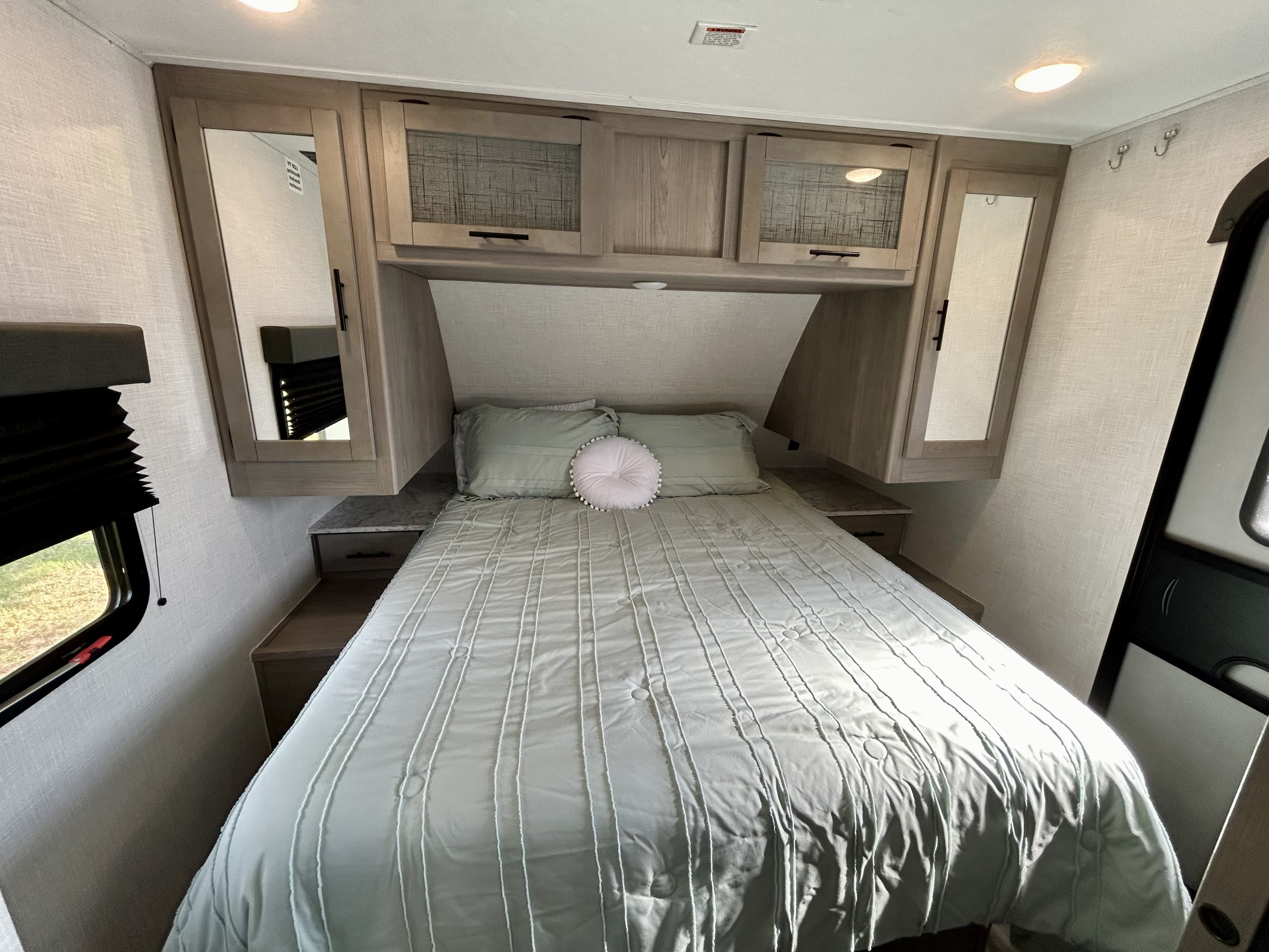 Clean & cozy RV rental bedroom features a comfortable bed with light green bedding, overhead cabinets, mirrors, and a window.. Coleman Dutchmen 2022
