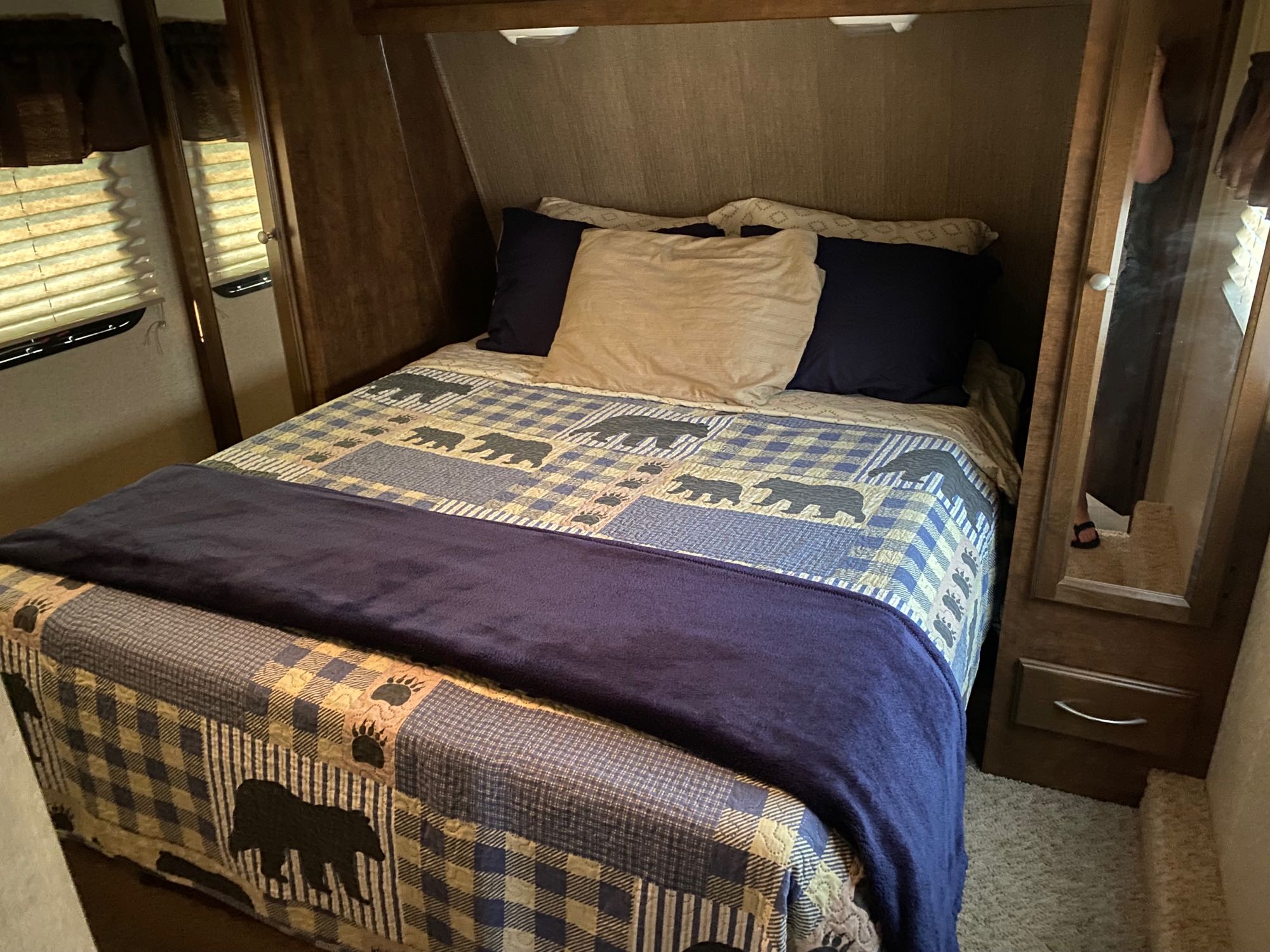 Inviting RV rental bedroom with a rustic bear quilt, blue blanket, pillows, and wood-toned walls. Includes mirrored closet and blinds.. Coachman Chaparral 2014