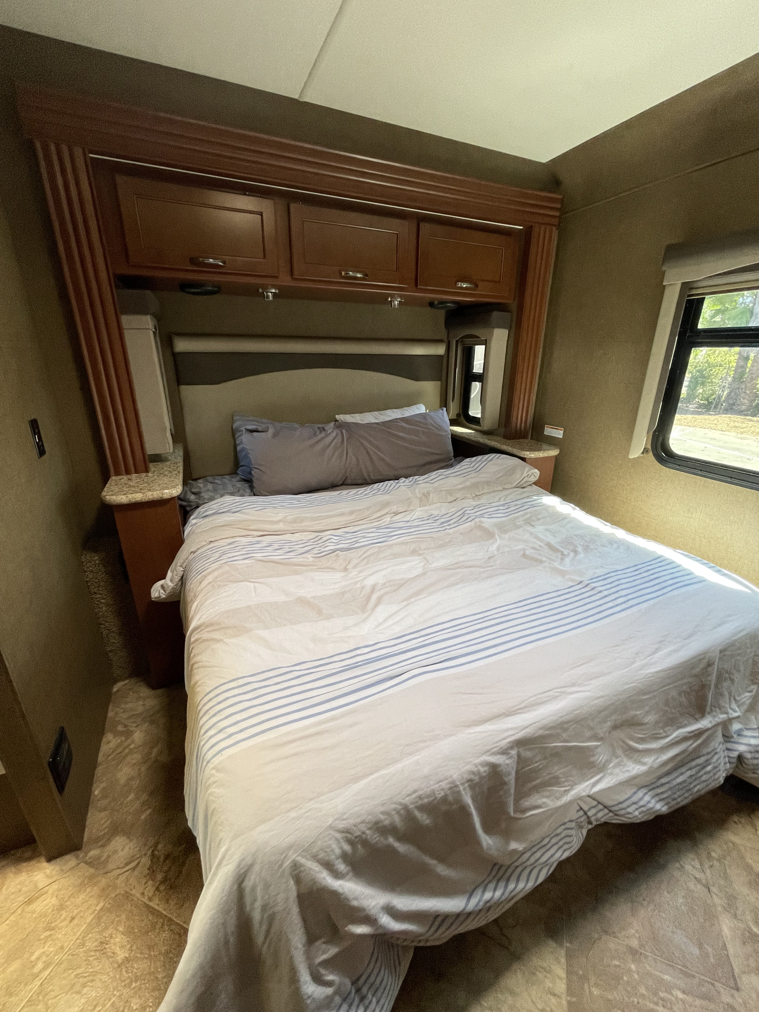 Cozy RV rental bedroom with a striped bed, wood cabinets, and a window showcasing the outdoors. Ideal for travel.. Thor Motor Coach A.C.E 2017