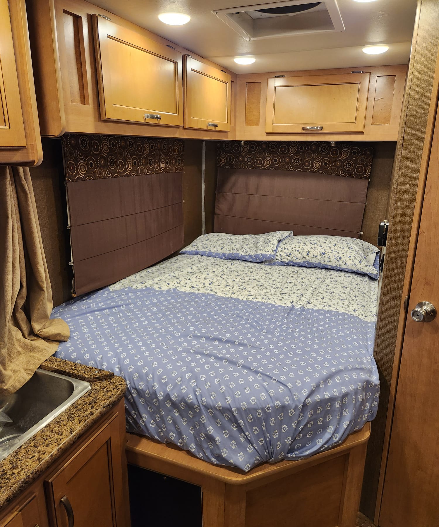 This compact RV rental bedroom features a cozy bed with blue and white bedding, overhead cabinets, and a small sink area.. Thor Four Winds 2017