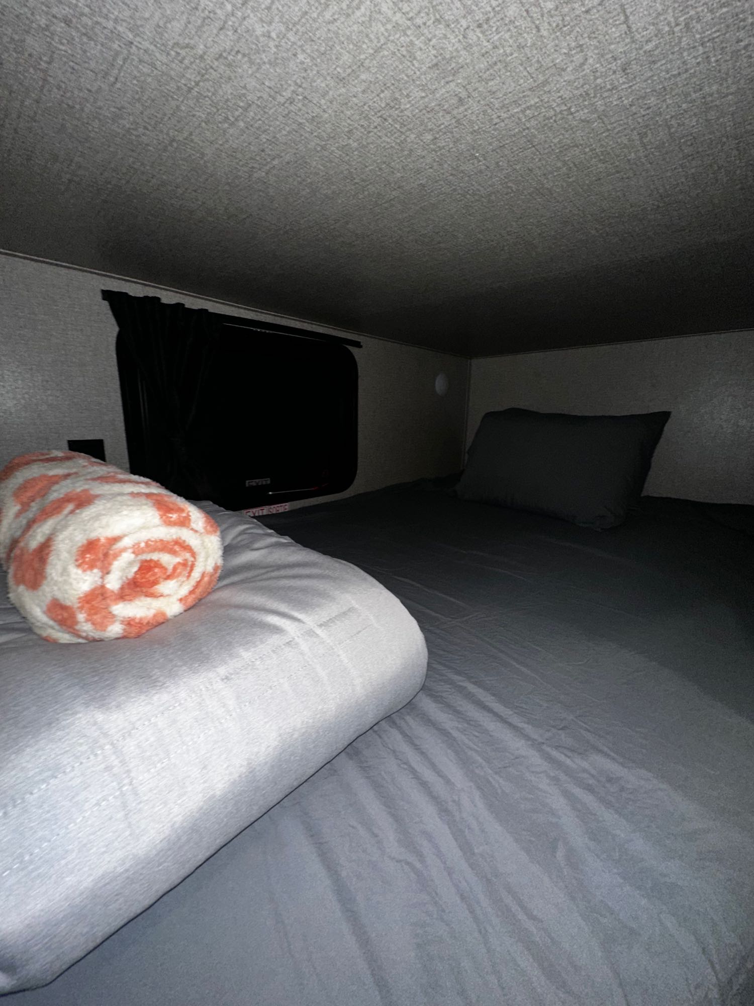 An RV rental's small, dark sleeping bunk features gray bedding, an orange/white blanket, and an exit window.. Jayco Jay Flight SLX Baja Edition 2021