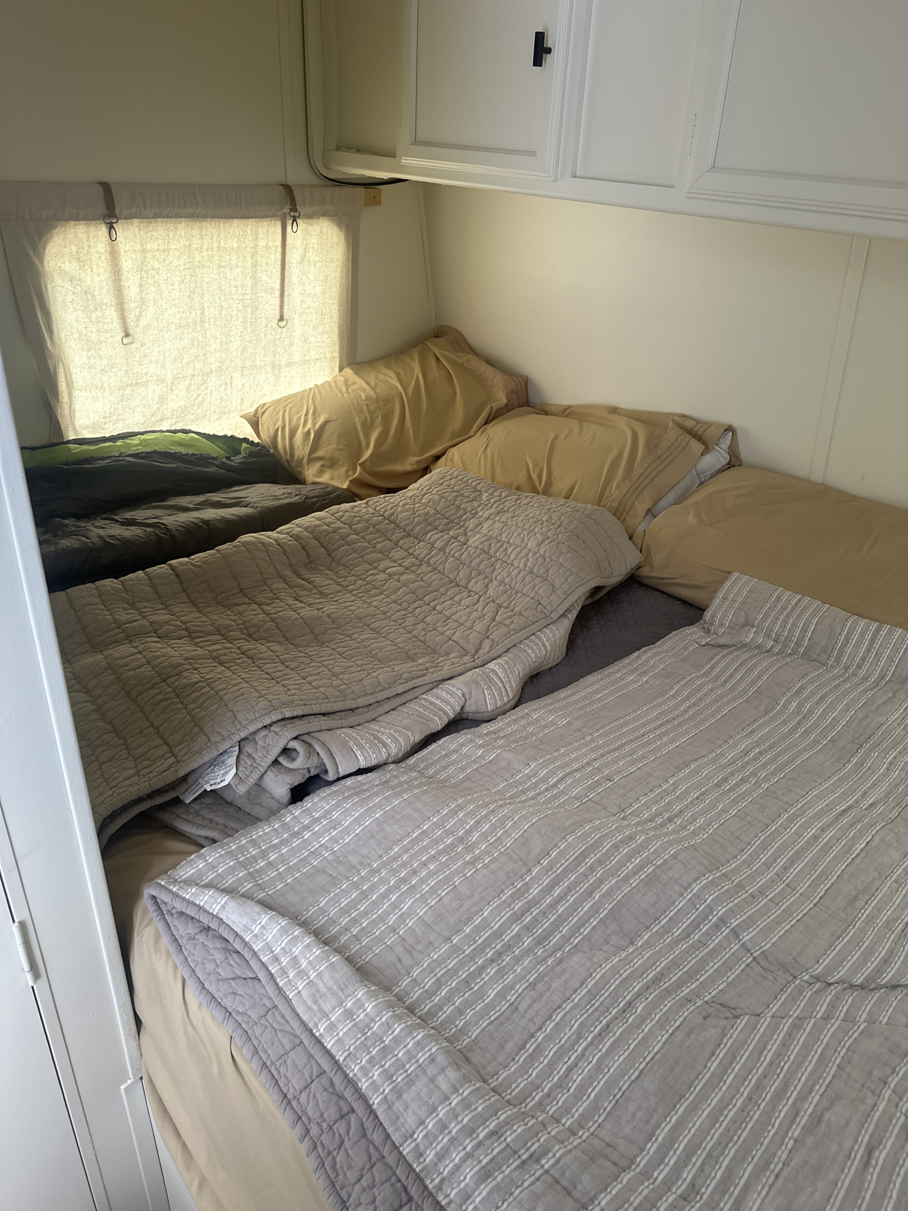 An RV rental's cozy bedroom, featuring multiple beds with comfy pillows, layered blankets, and natural light from a curtained window.. Skyline Nomad 2008