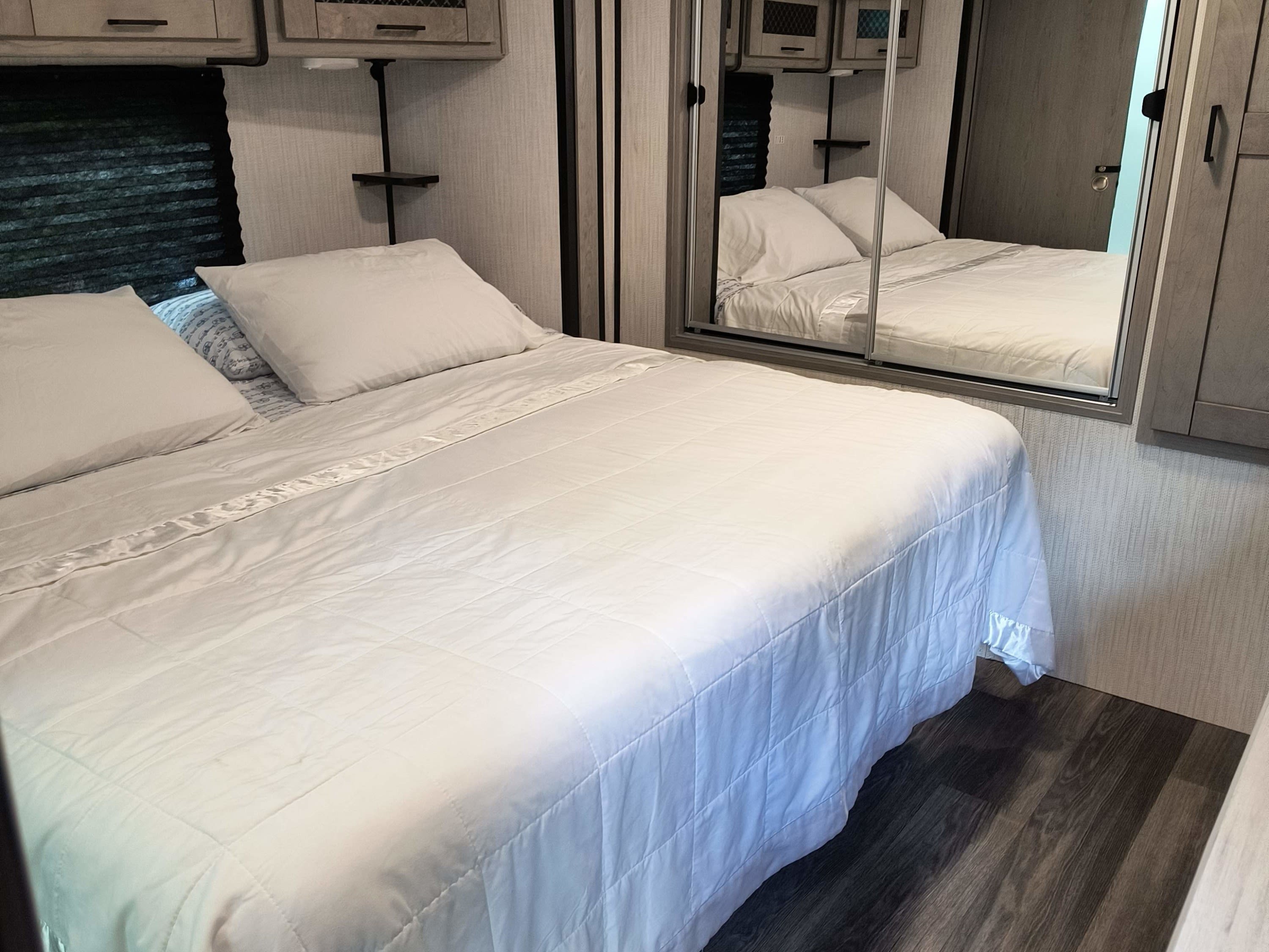 Spacious RV rental bedroom with crisp white bedding, overhead storage, and mirrored closet doors. Dark wood flooring.. Heartland RVs Mallard 2021