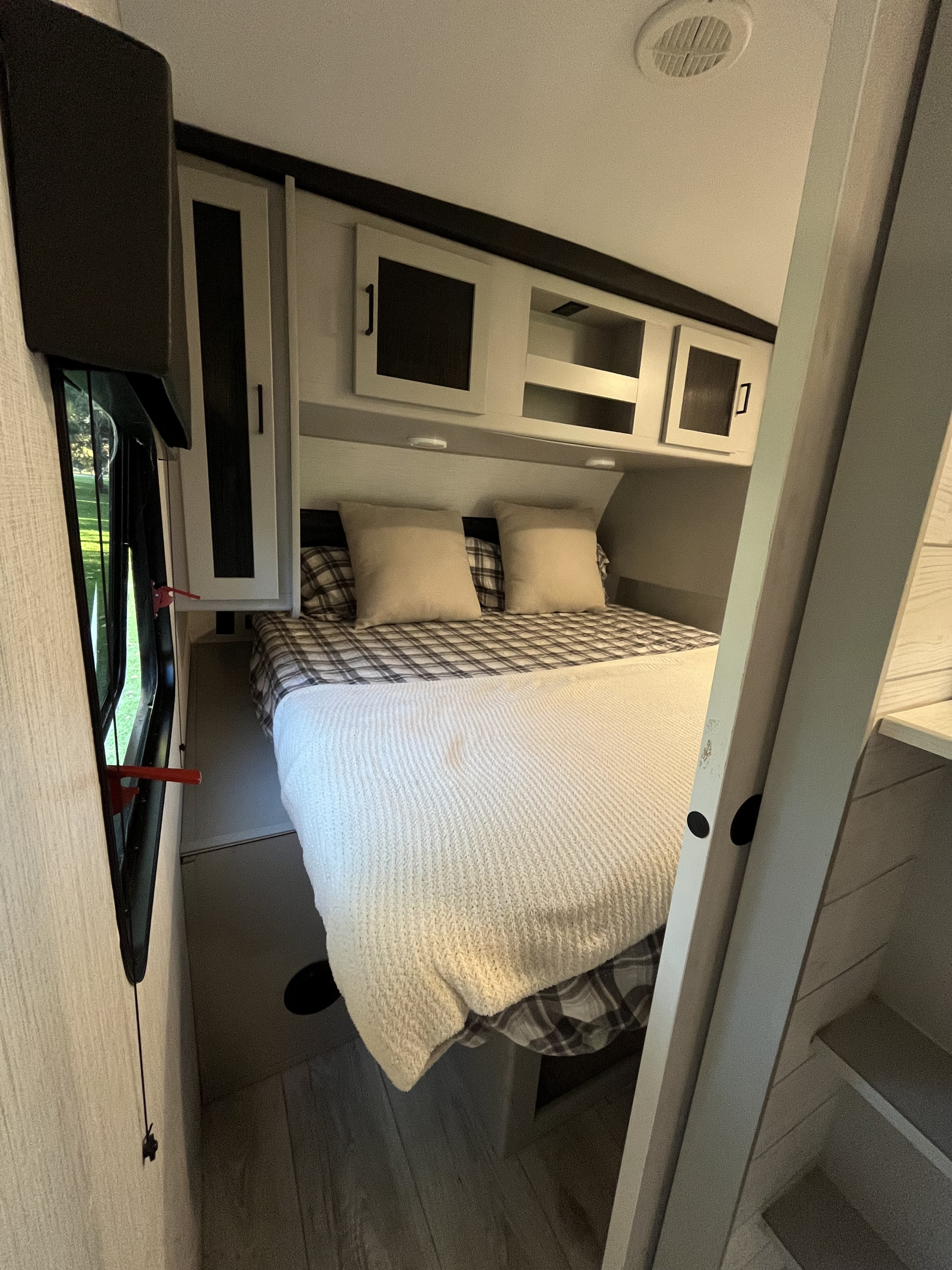This RV rental bedroom offers a cozy bed with plaid bedding, overhead cabinets, and a window for scenic views.. Heartland RVs North Trail 2022