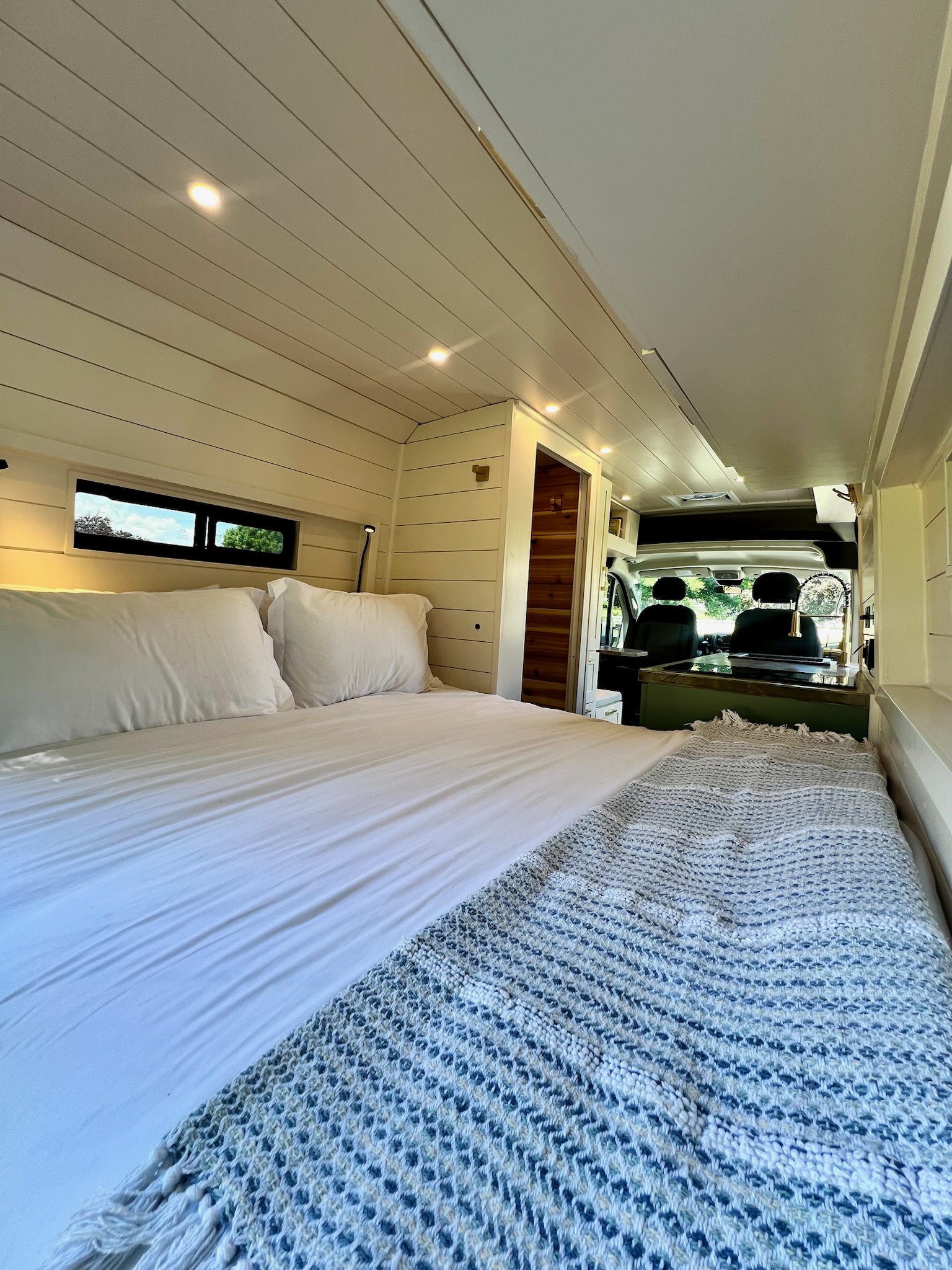 Spacious rv rental interior. White shiplap walls & ceiling, cozy bed with white sheets & blue blanket, kitchenette visible.. Custom by Nothing Ventured Vans 2021