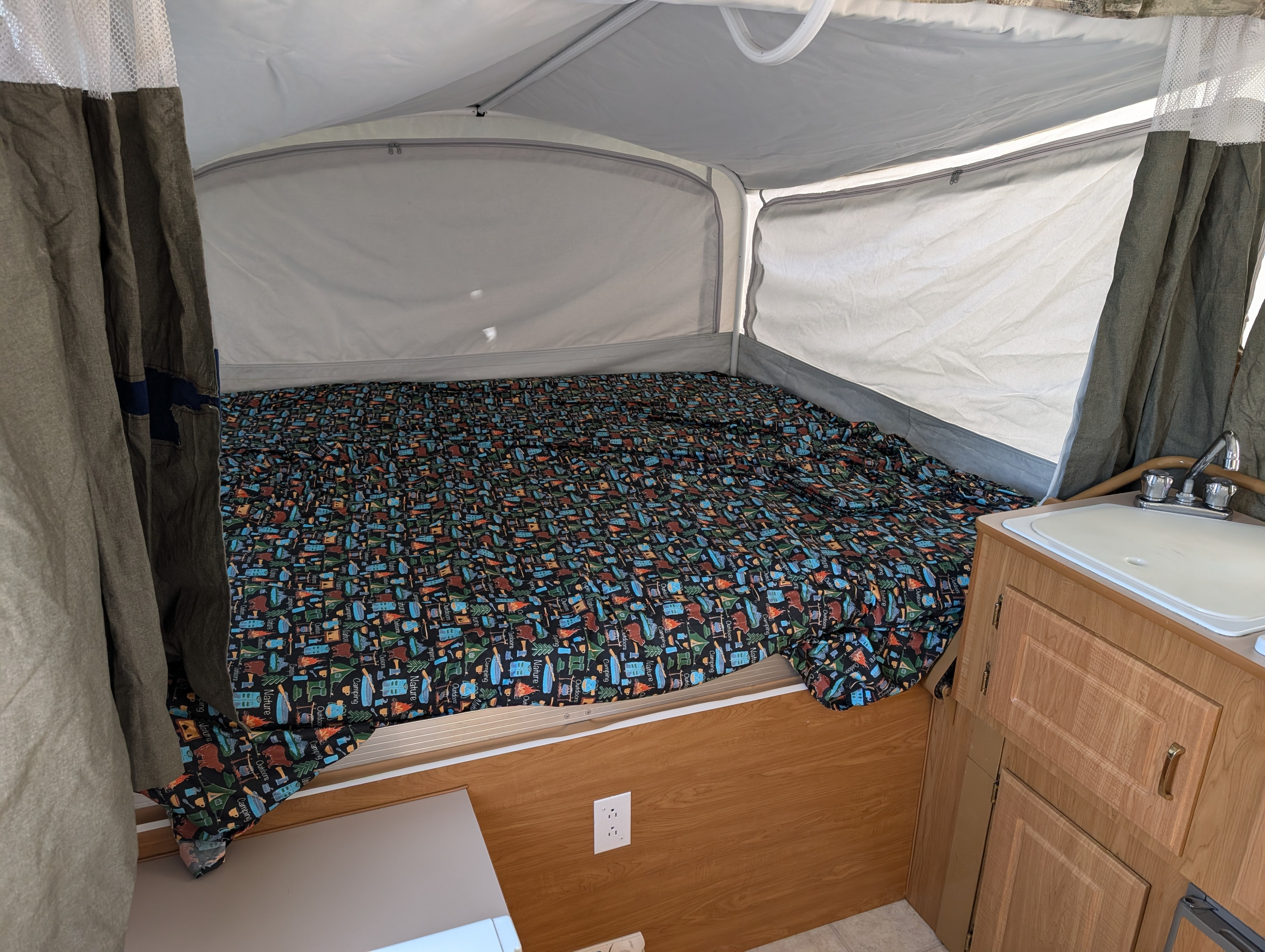 Cozy RV rental pop-up camper interior featuring a large bed with a camping-themed blanket and a compact kitchenette with a sink.. Coleman Utah 2003