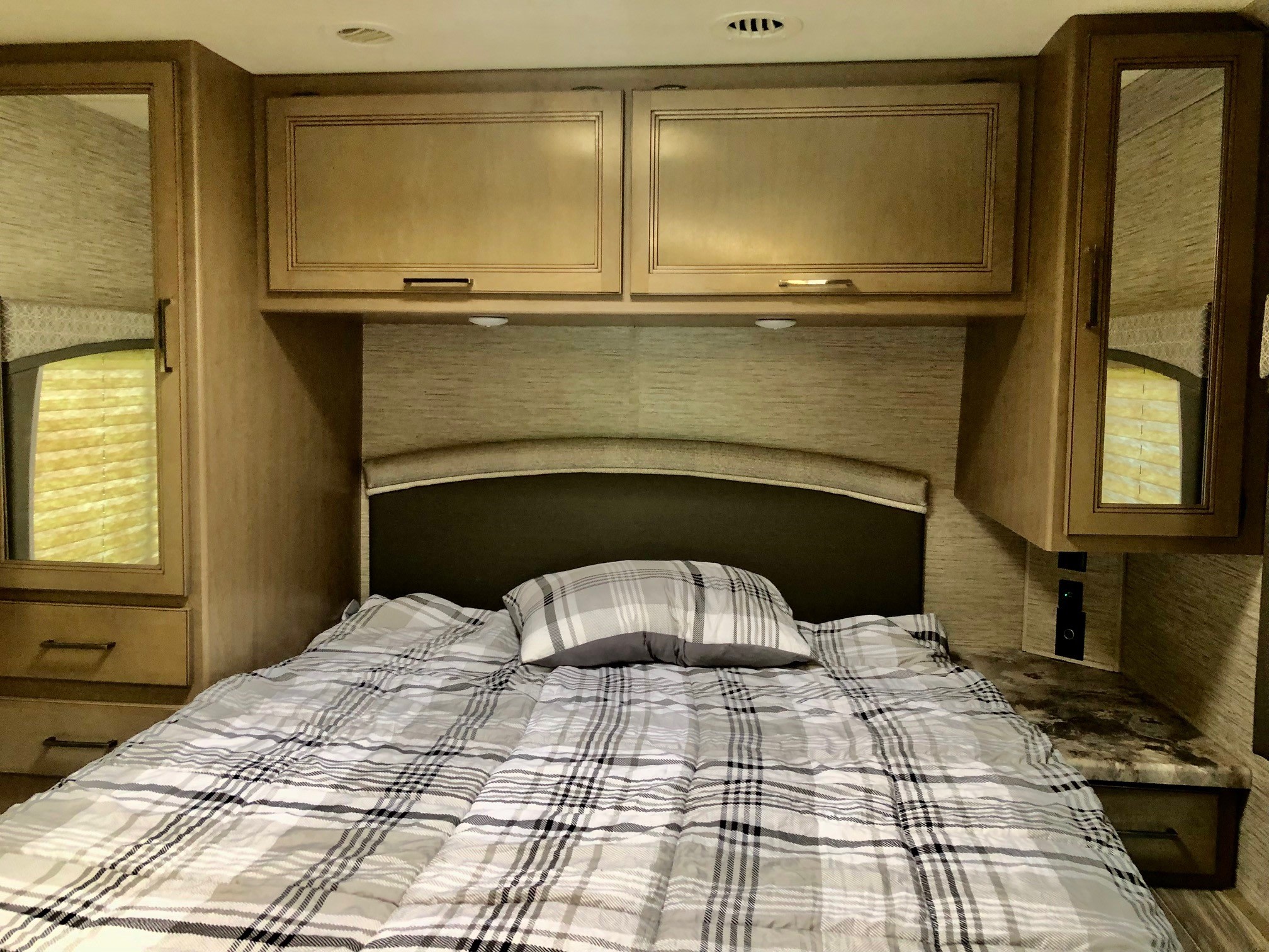 An RV rental bedroom features a comfy bed with plaid bedding, overhead storage, and mirrored cabinets on each side.. Thor Motor Coach Four Winds 2023