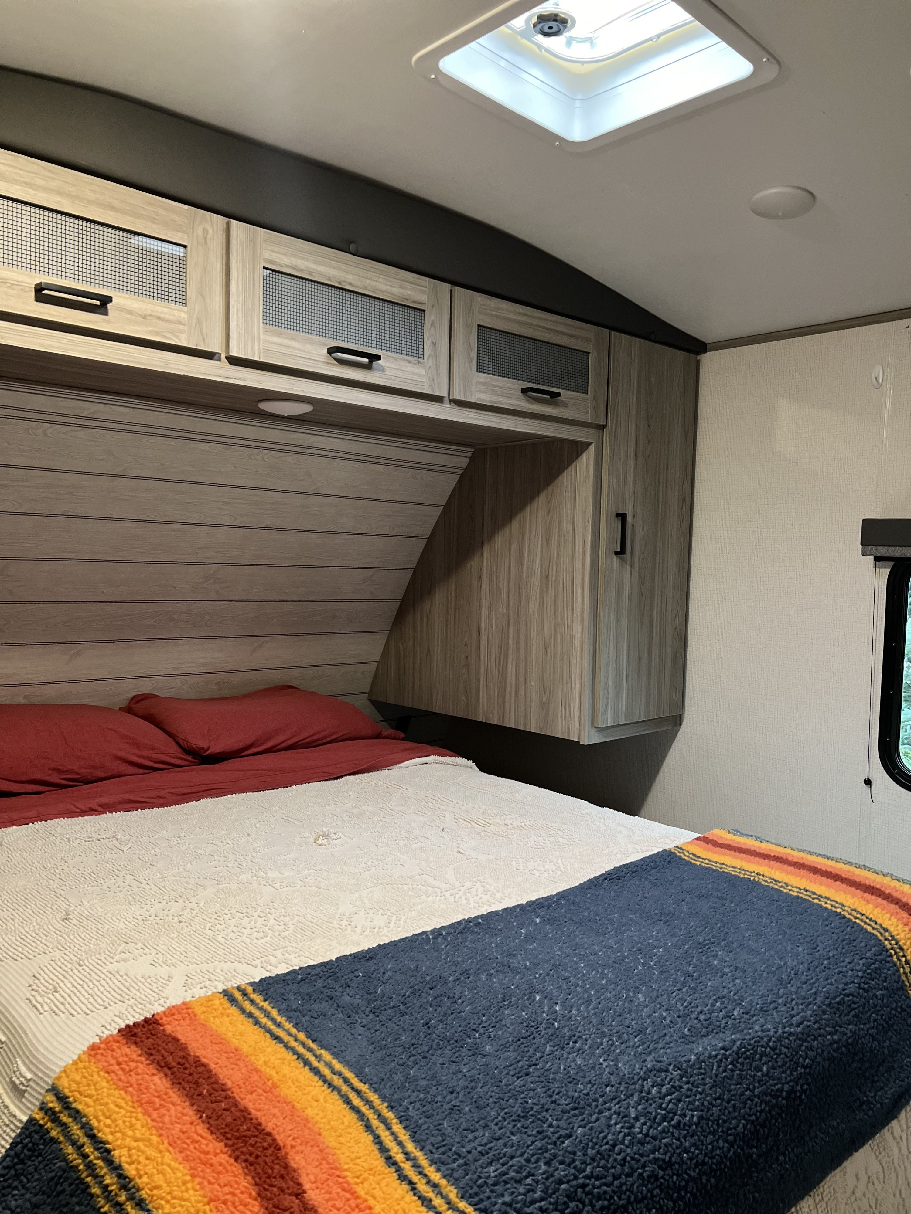 Cozy RV rental bedroom with light wood cabinets, striped blanket, red pillows, and overhead vent. Perfect for a comfy road trip.. Keystone RV Passport GT Ultra Lite 2023