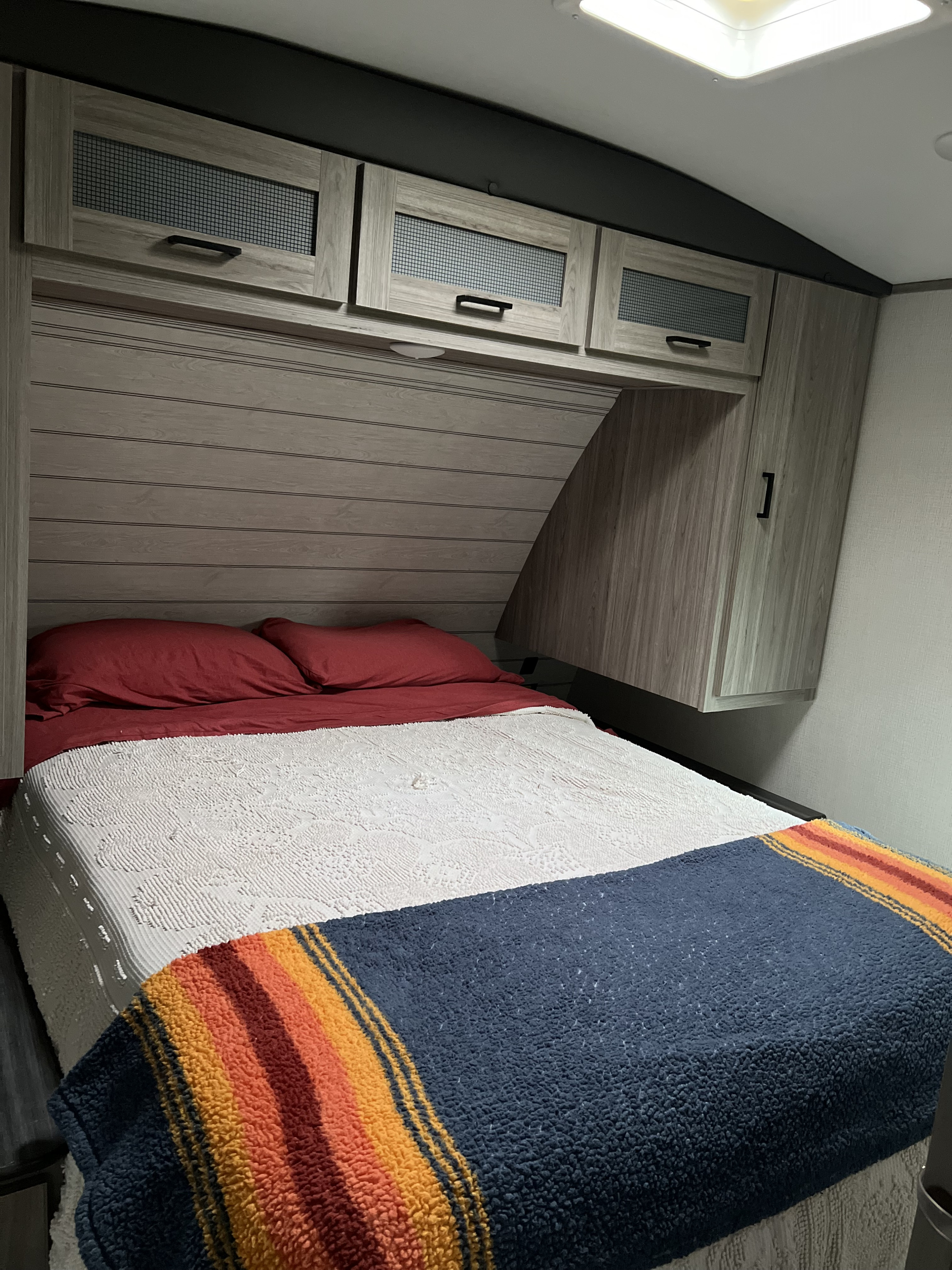 Cozy RV rental bedroom features a bed with red pillows, white comforter, striped blanket, and overhead wood cabinets.. Keystone RV Passport GT Ultra Lite 2023