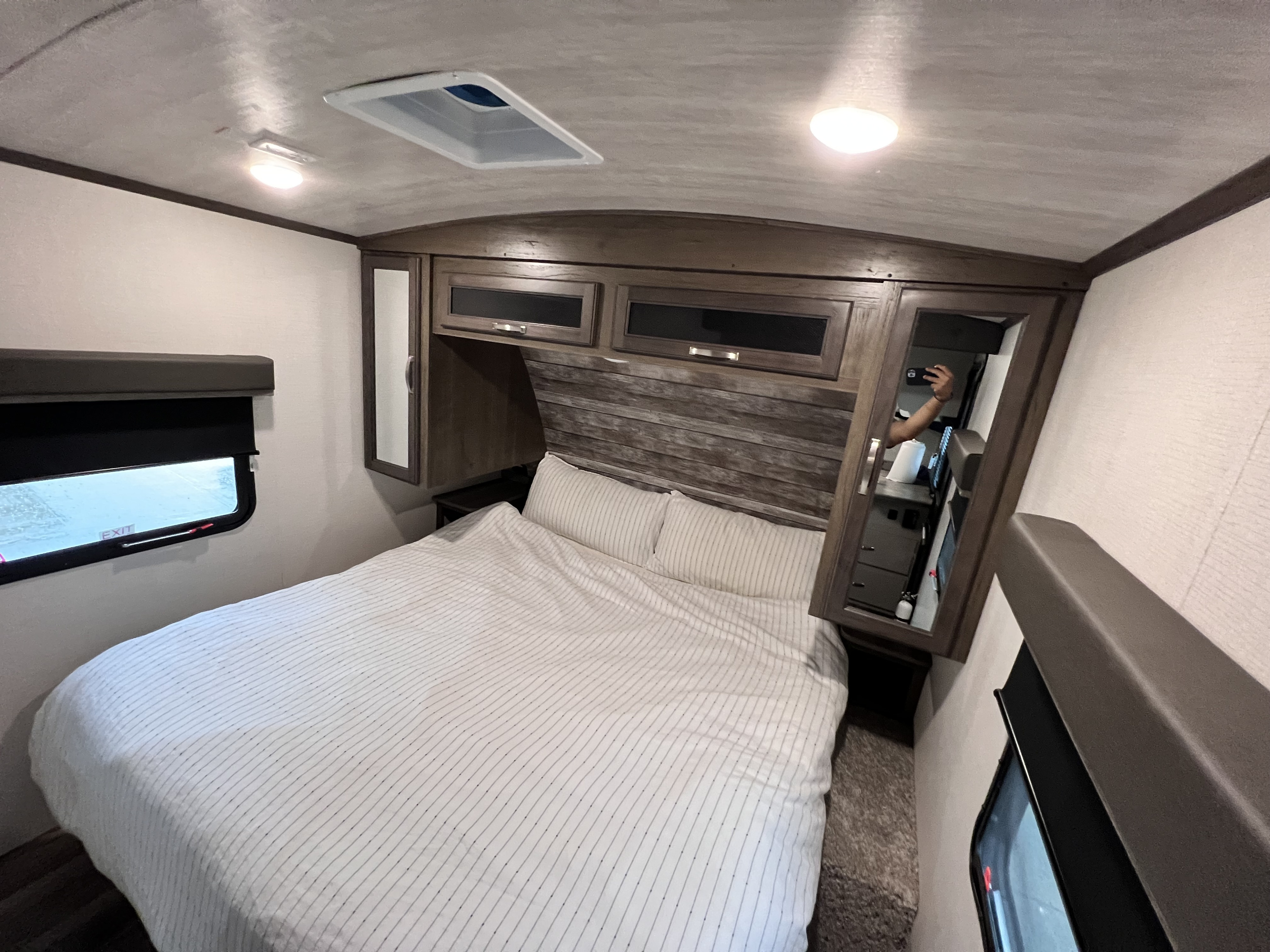 RV rental bedroom with striped bed, overhead wood cabinets, and an emergency exit window. Includes ceiling lights and vent.. Keystone RV Cougar 2022