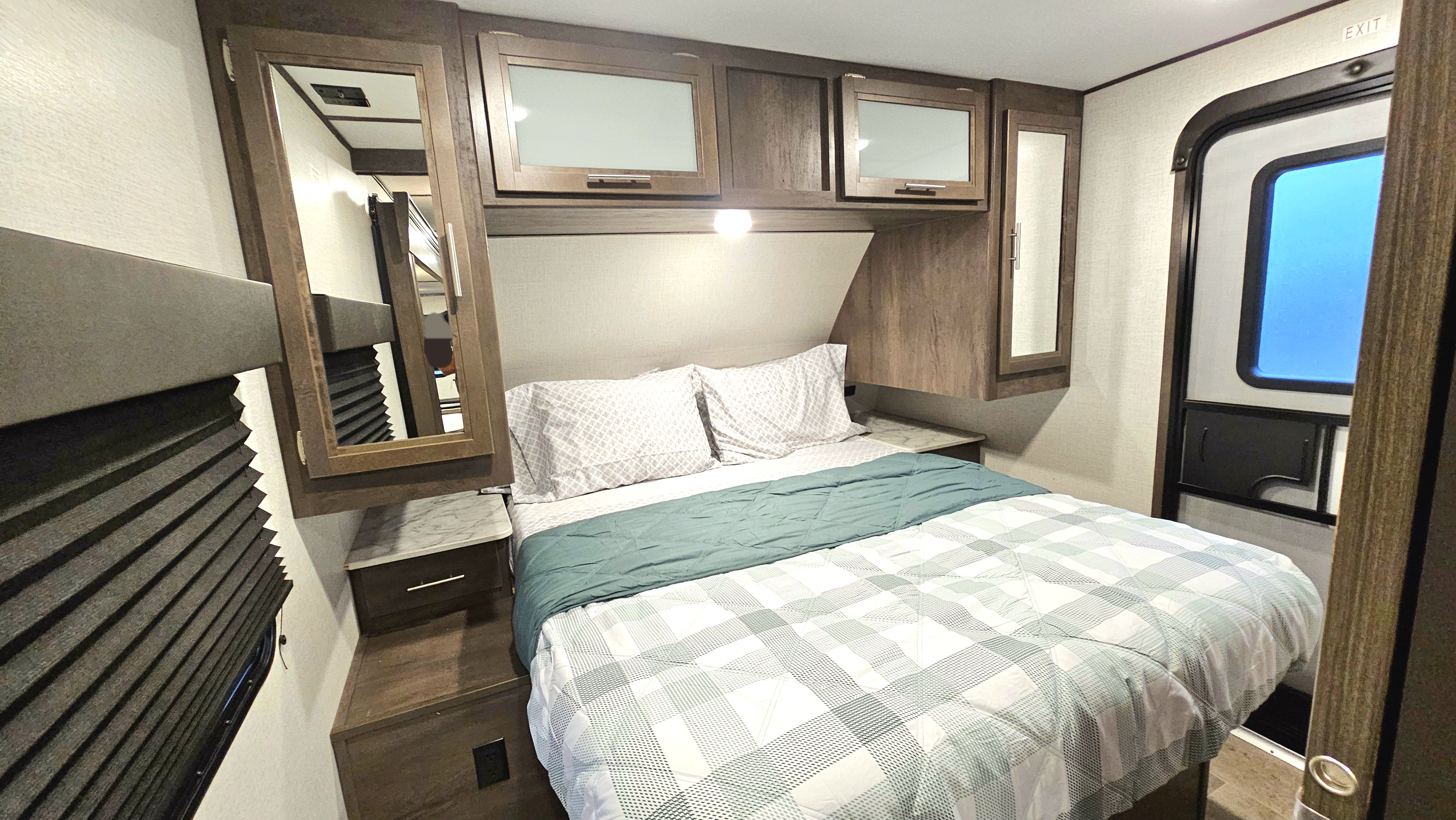 RV rental bedroom featuring a patterned bed, wood-look cabinets, nightstands, and a side exit door.. Dutchmen Coleman Light 2021
