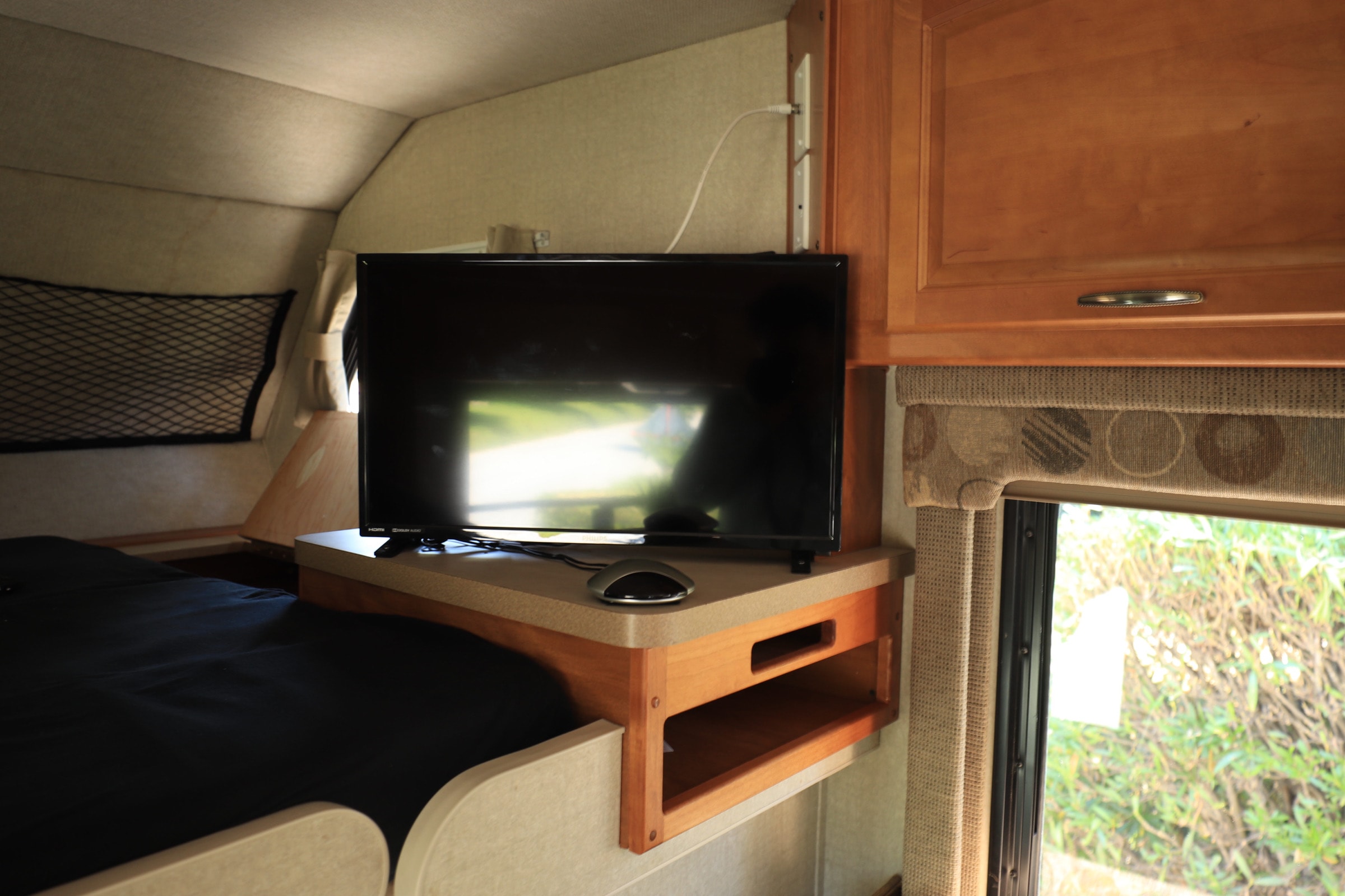 Inside an RV rental, a compact sleeping area features a dark bed, TV on a shelf, and a window displaying green foliage.. Fleetwood Tioga 31m 2007