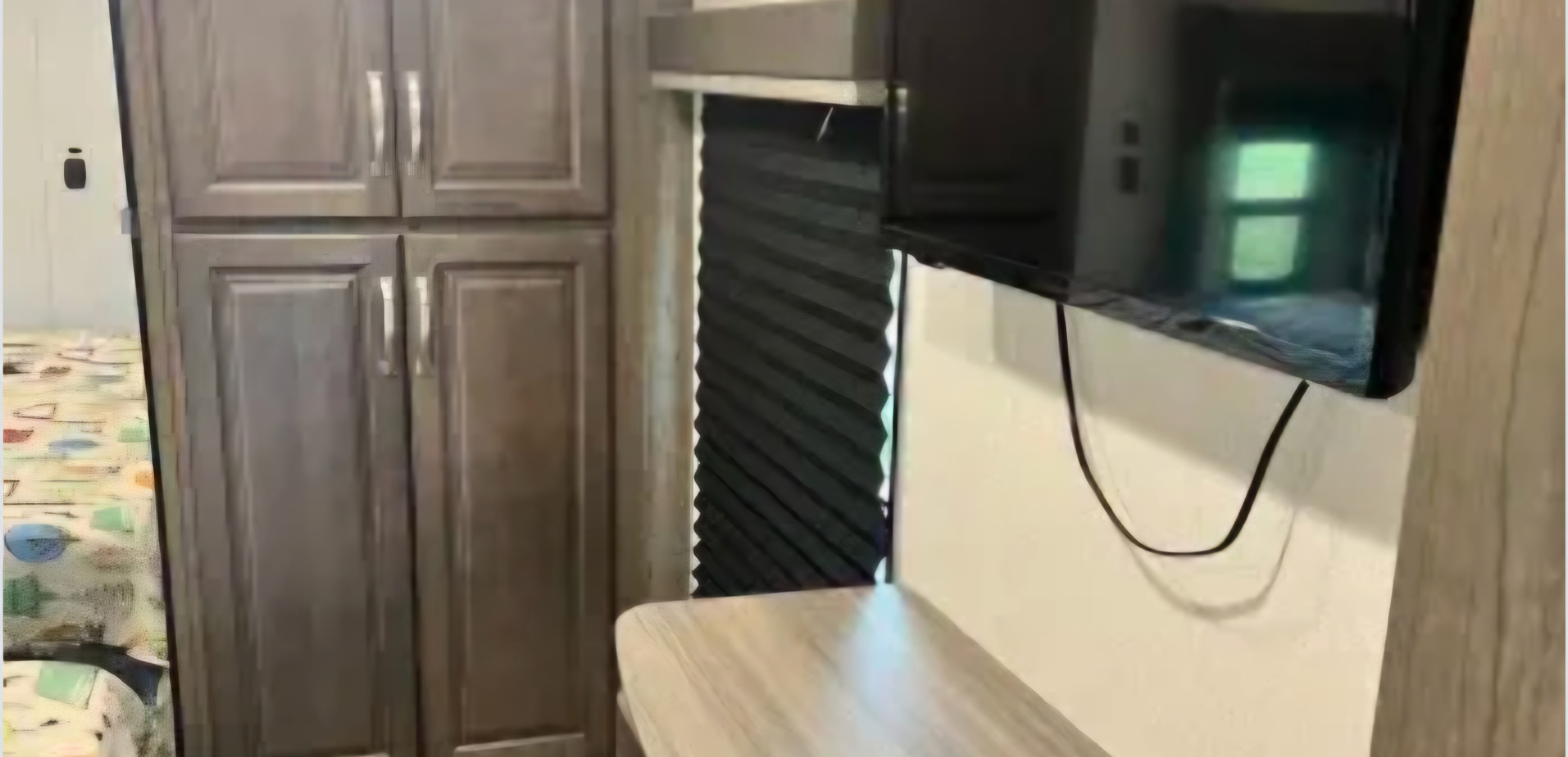 RV rental interior: dark wood cabinets, a mounted TV with cord, pleated window blinds, and a light counter space. A bed is on the left.. Keystone RV Cougar 2021