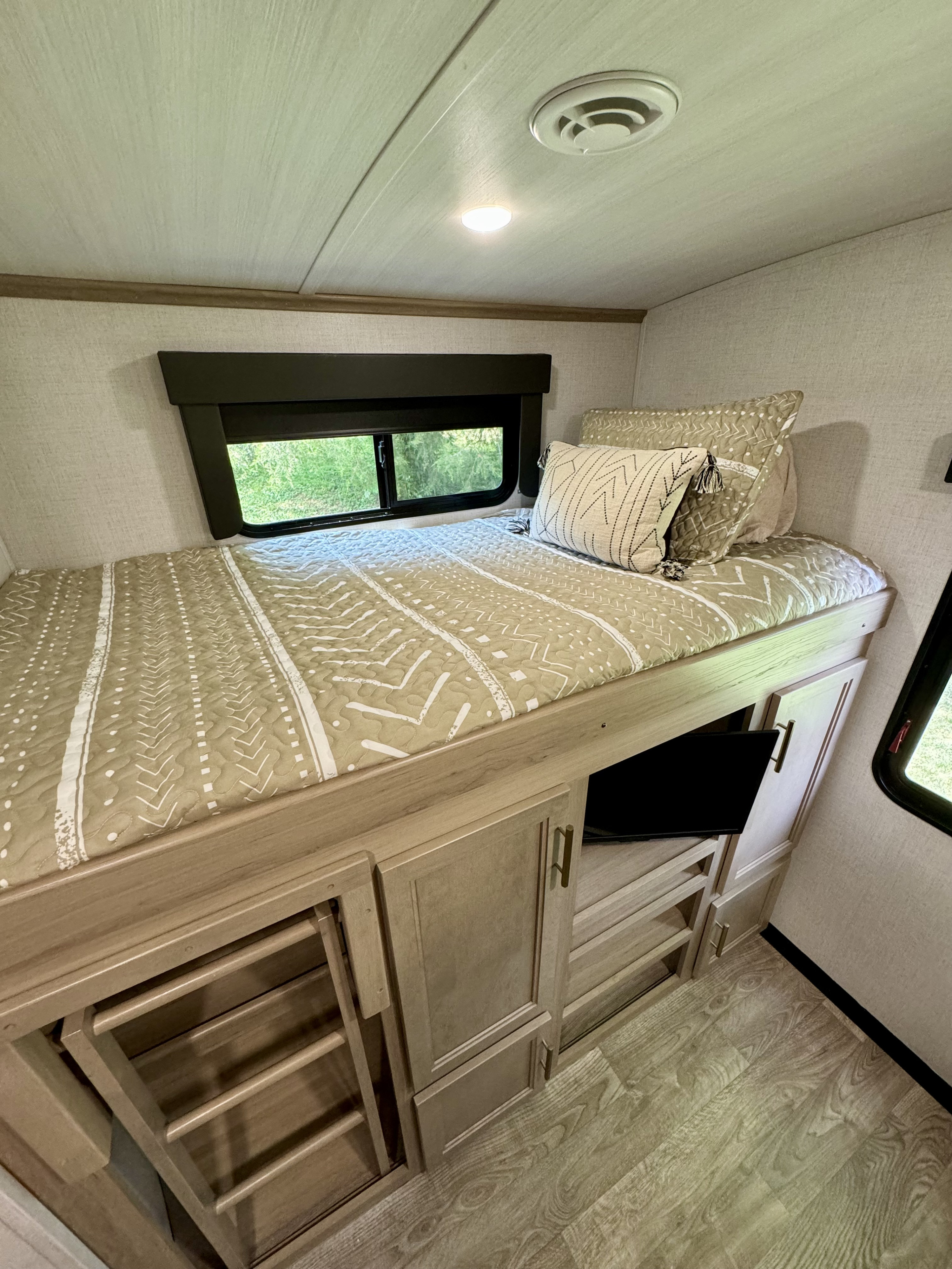 An inviting RV rental interior: a raised bed with light bedding, built-in storage, ladder, TV, and window view.. Keystone RV 340BH 2024