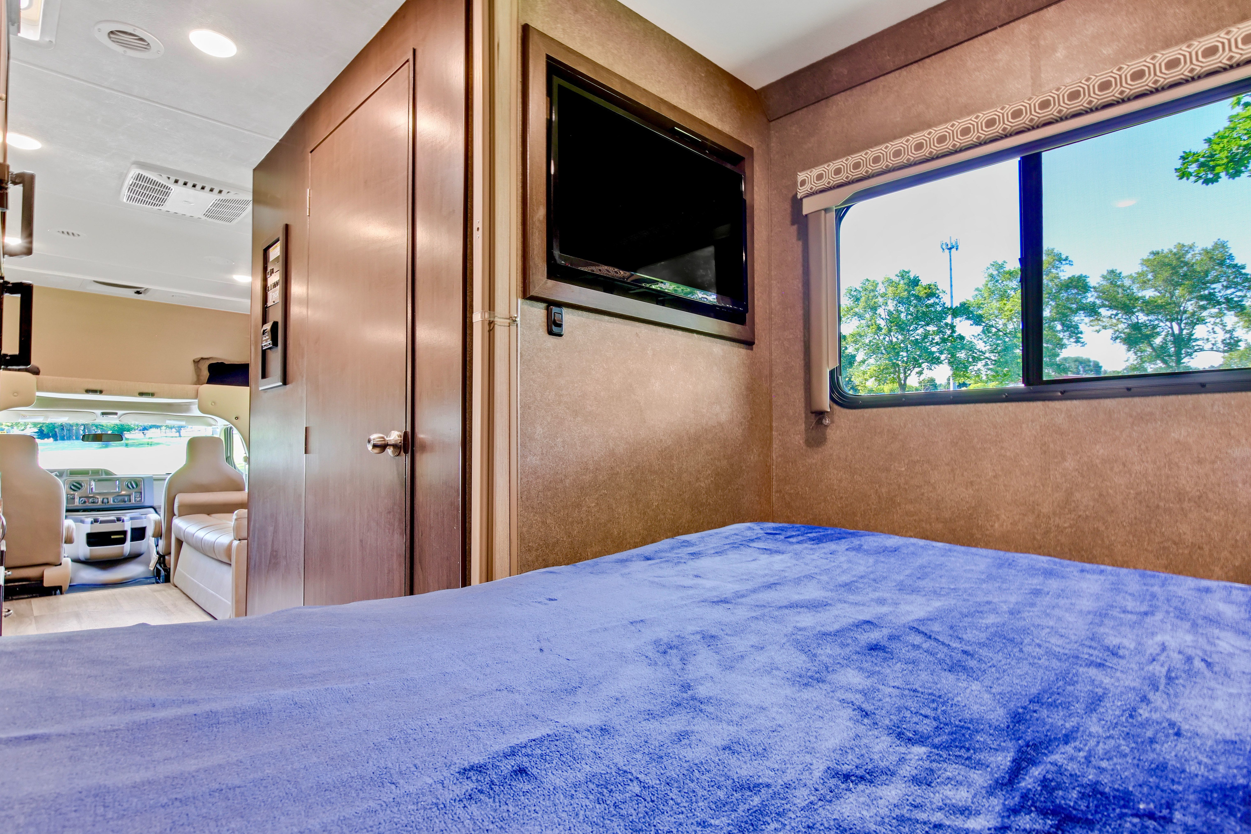 Inside this rv rental bedroom: a comfy bed with blue blanket, wall-mounted TV, window overlooking trees, and front cabin.. Thor Motor Coach Four Winds 2020