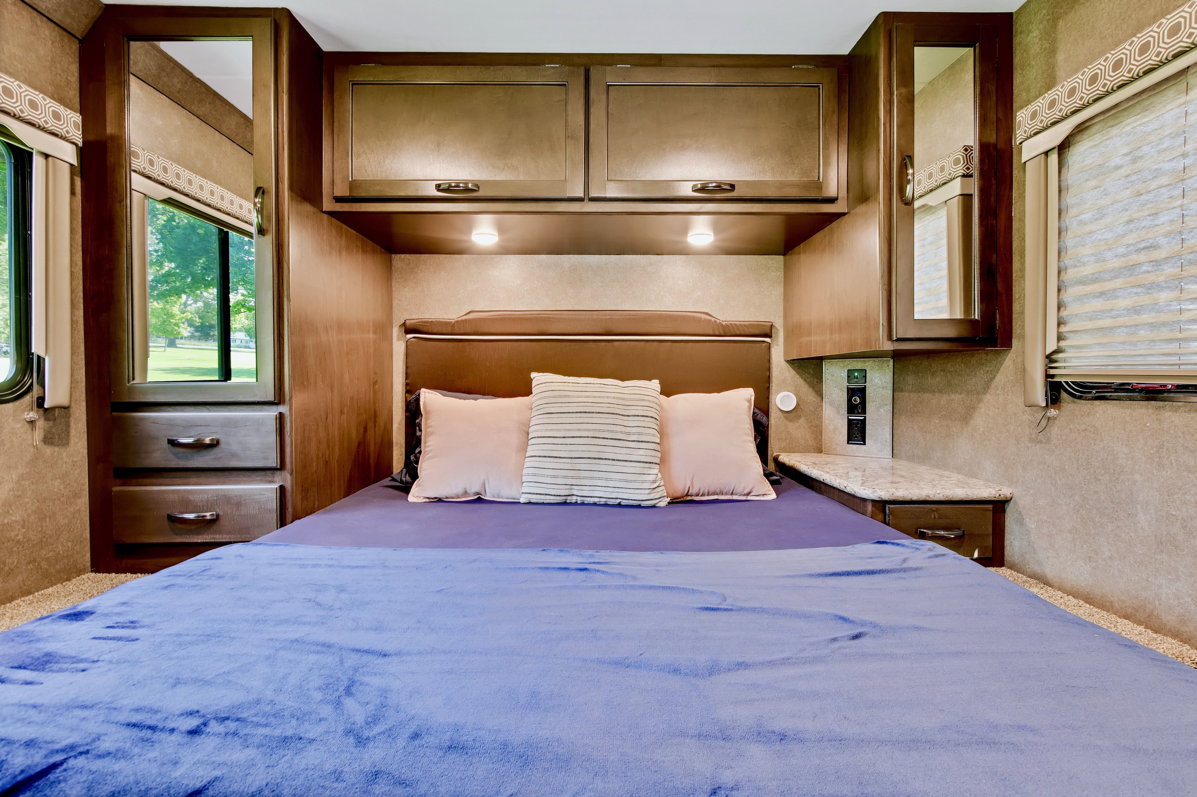 Inviting RV rental bedroom with a blue blanketed bed, overhead storage, and side windows showcasing outdoor greenery.. Thor Motor Coach Four Winds 2020