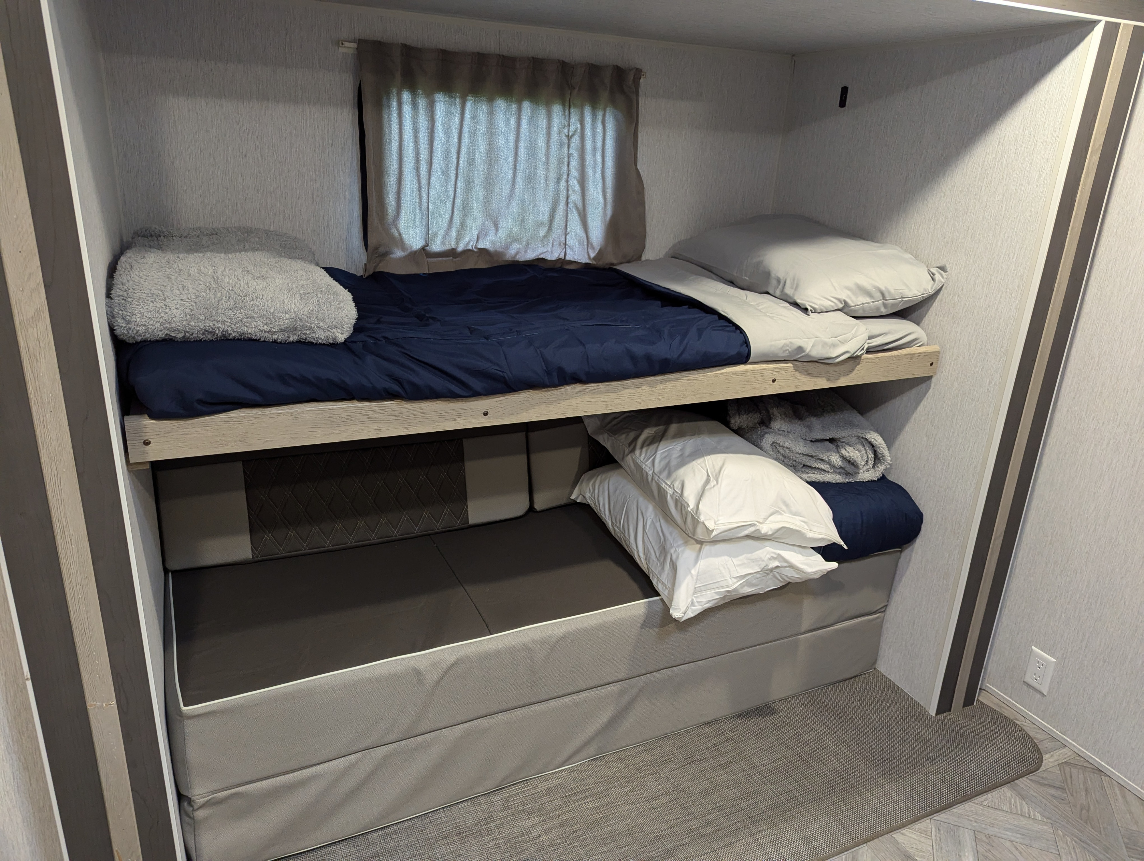 This compact RV rental features cozy bunk beds. The top bunk has a blue comforter; the lower has gray cushions with white pillows.. Forest River Wildwood 2023