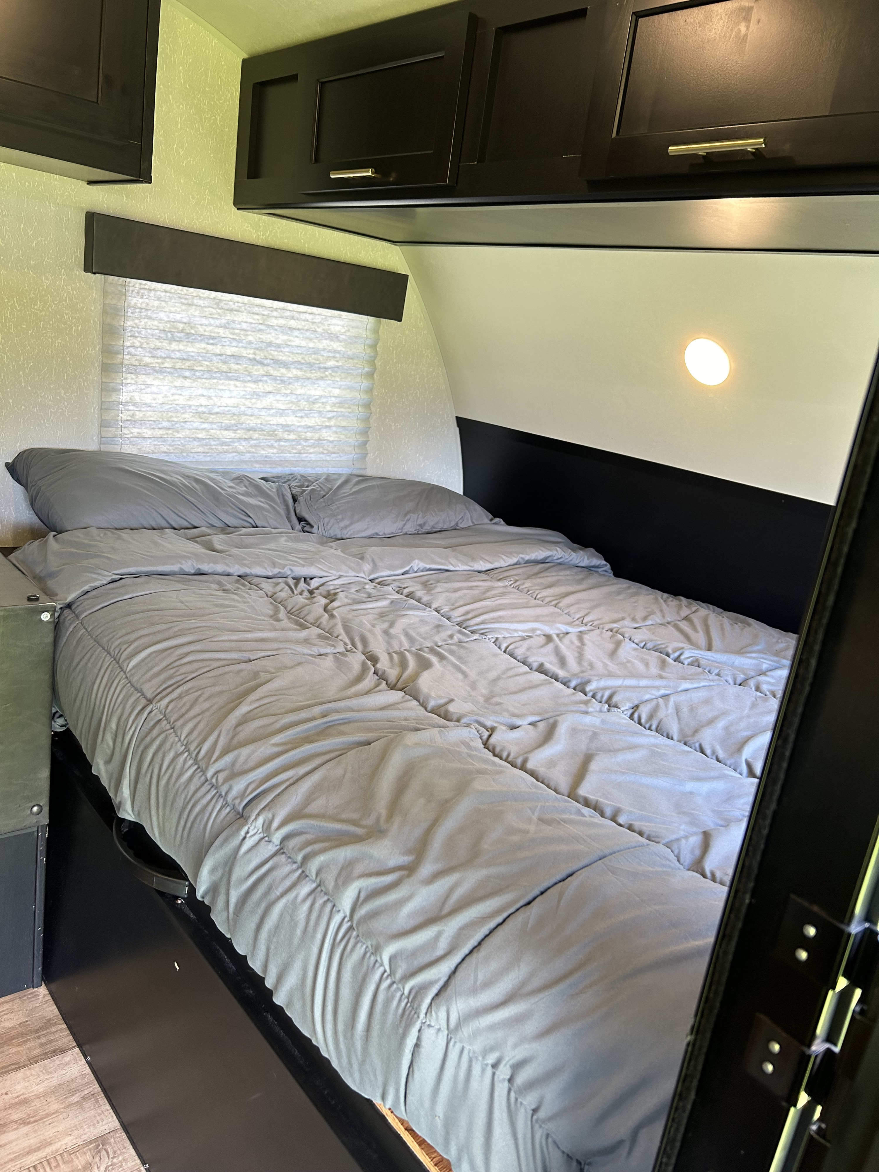 Cozy RV rental bedroom features a grey bed, window with blinds, overhead cabinets, and a wall light.. Forest River Cherokee Wolf Pup 2022
